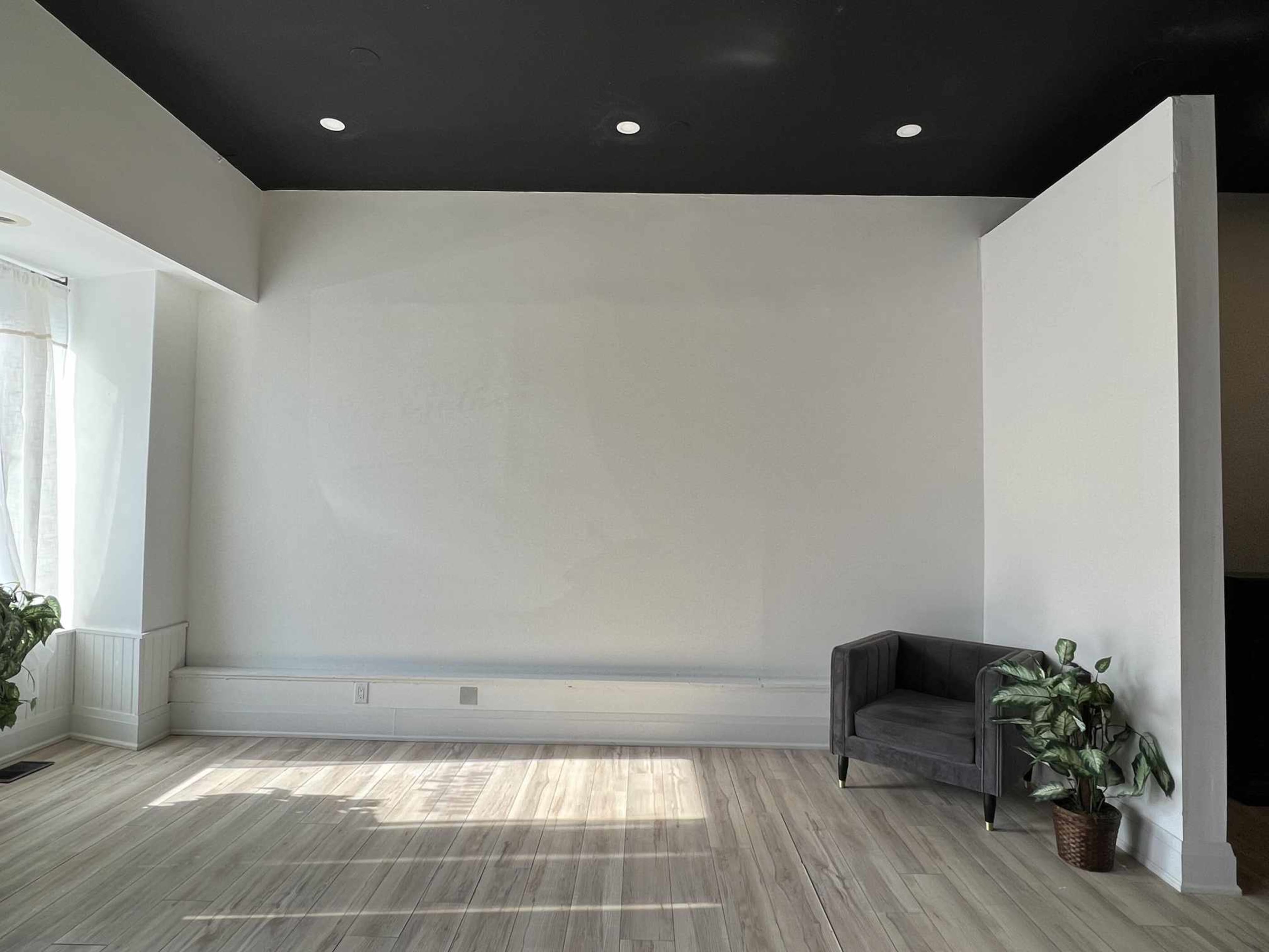 A sparsely furnished room with a gray chair and a potted plant in one corner, featuring light-colored wood flooring and white walls.
