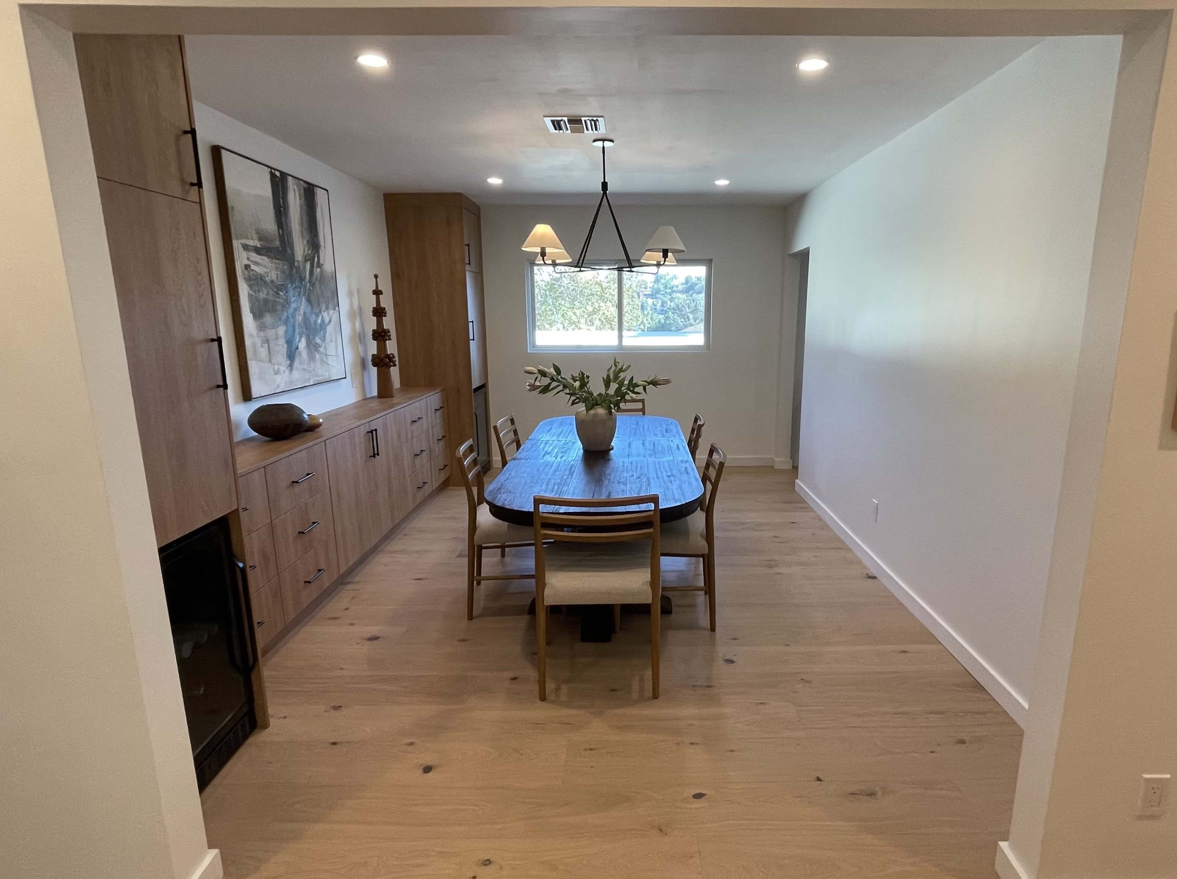A dining room features a wooden table surrounded by chairs, with a sideboard and artwork visible on the walls.