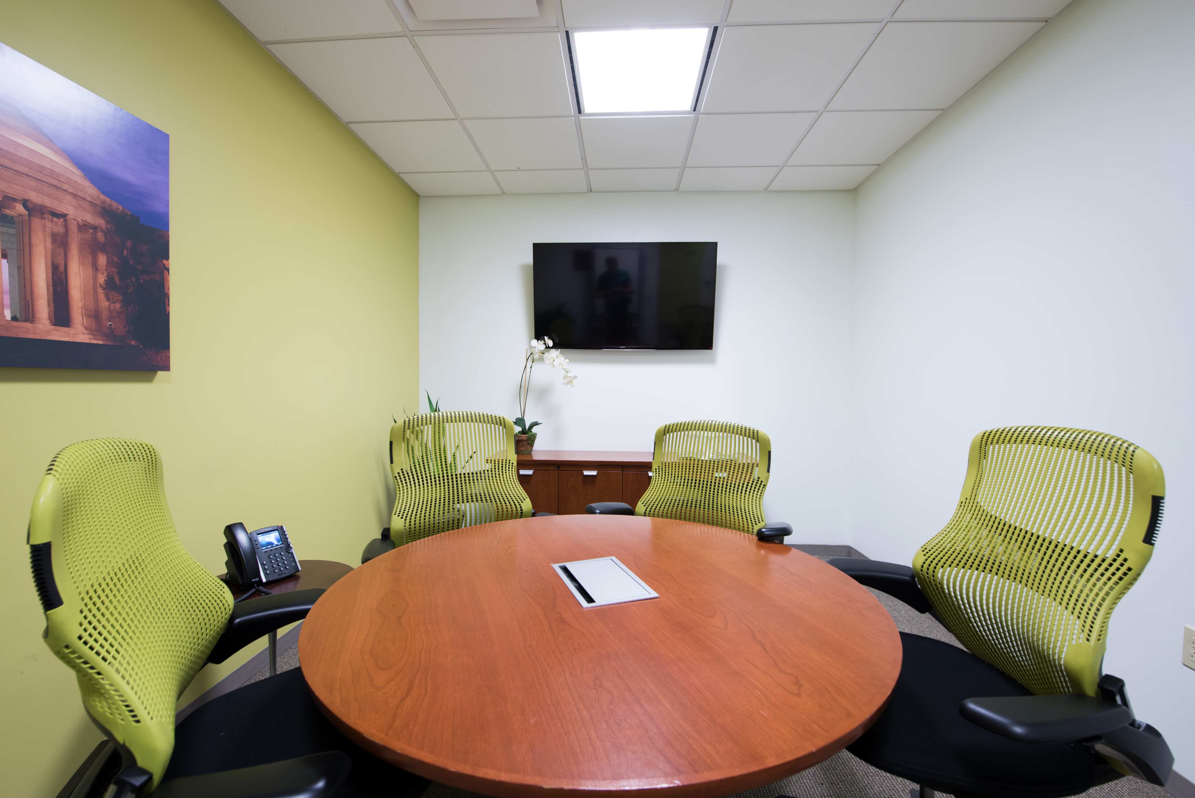 A meeting room features a round wooden table surrounded by four green office chairs, a television mounted on the wall, and a phone on a side table.