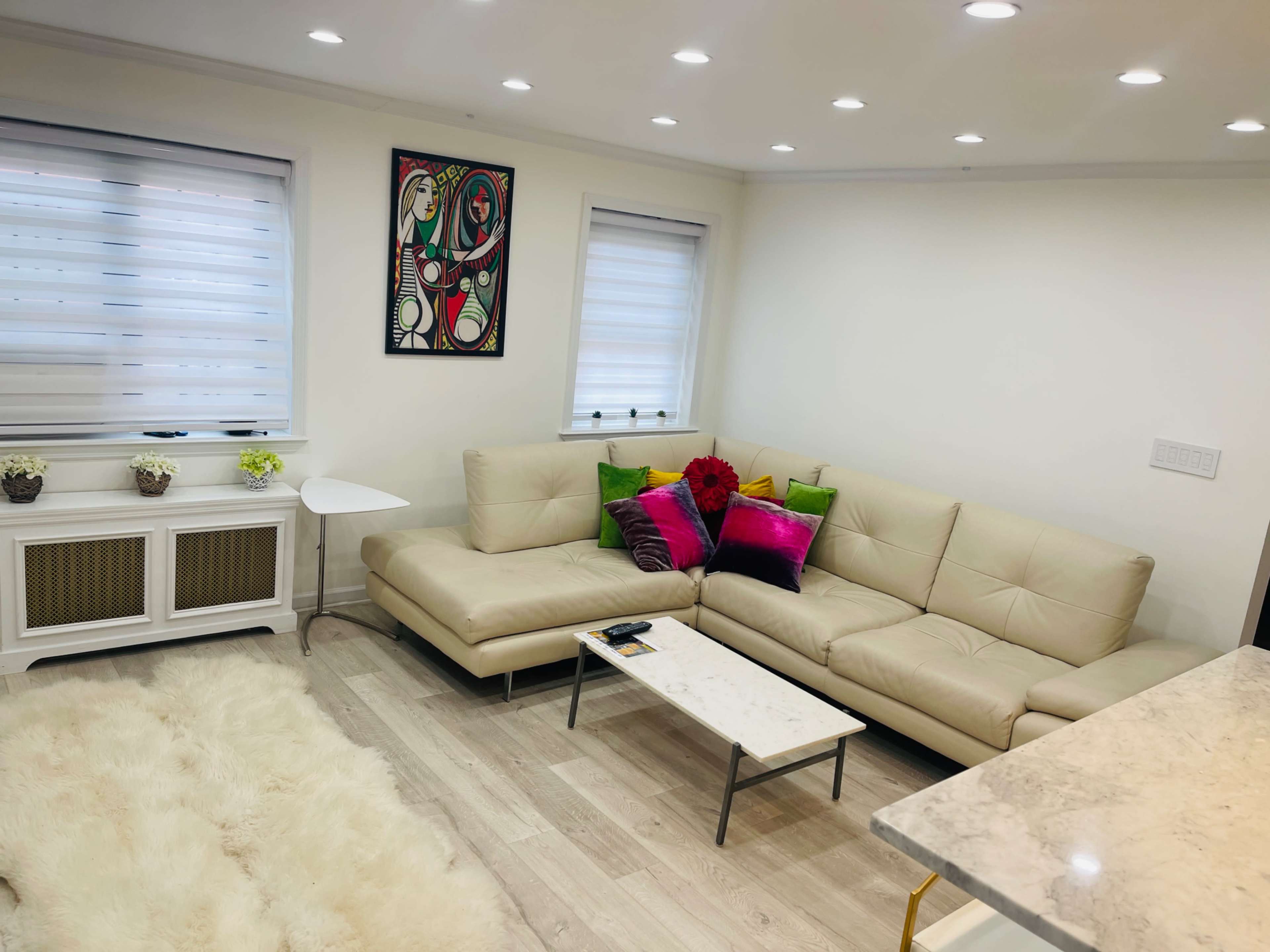 A modern living room featuring a beige leather sectional sofa with colorful cushions, a small white side table, and a marble-topped coffee table atop a light-colored wooden floor.