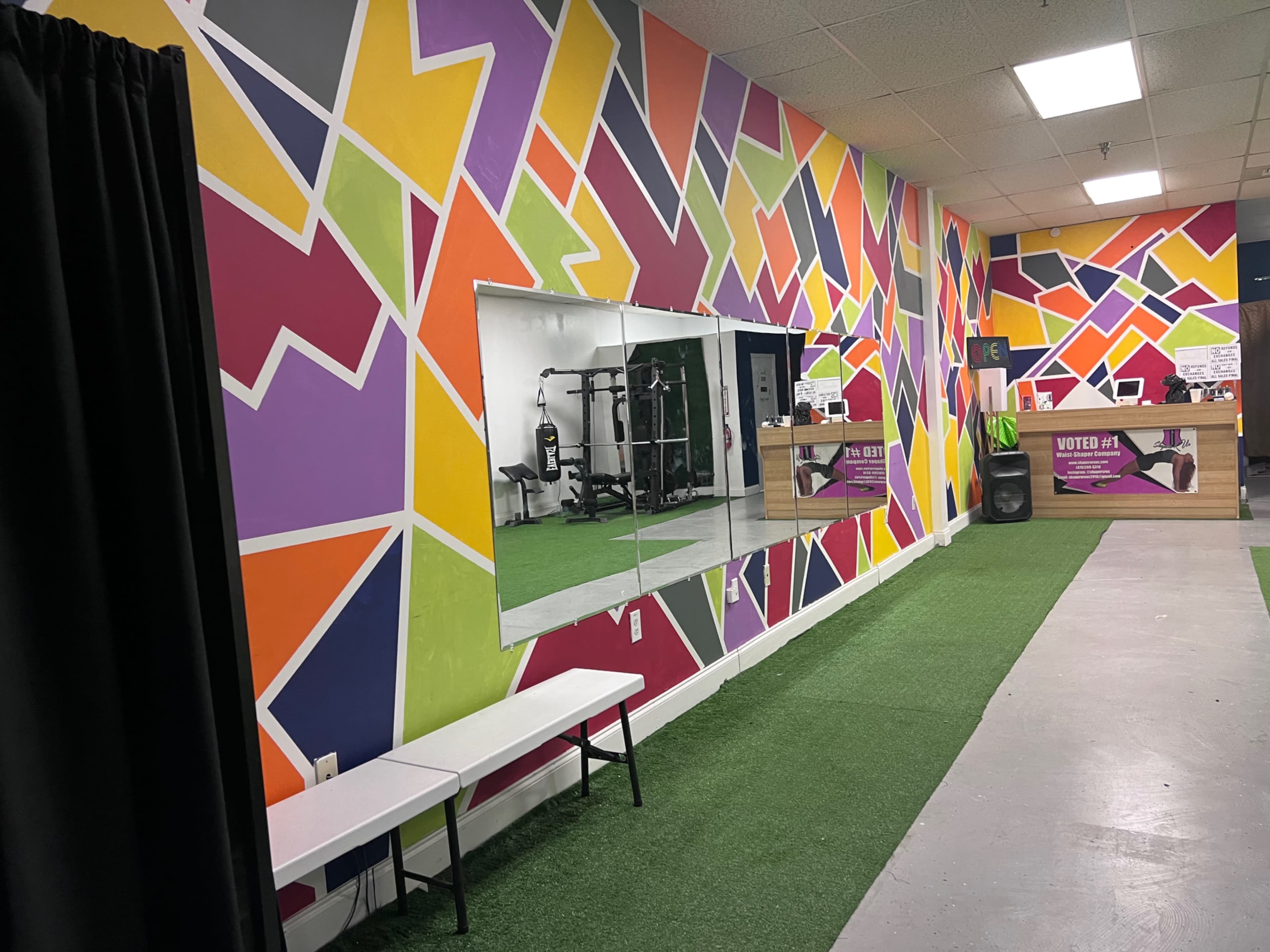A vibrant gym interior with a colorful geometric mural on the walls, a mirror, a green turf area, and workout equipment visible in the background.