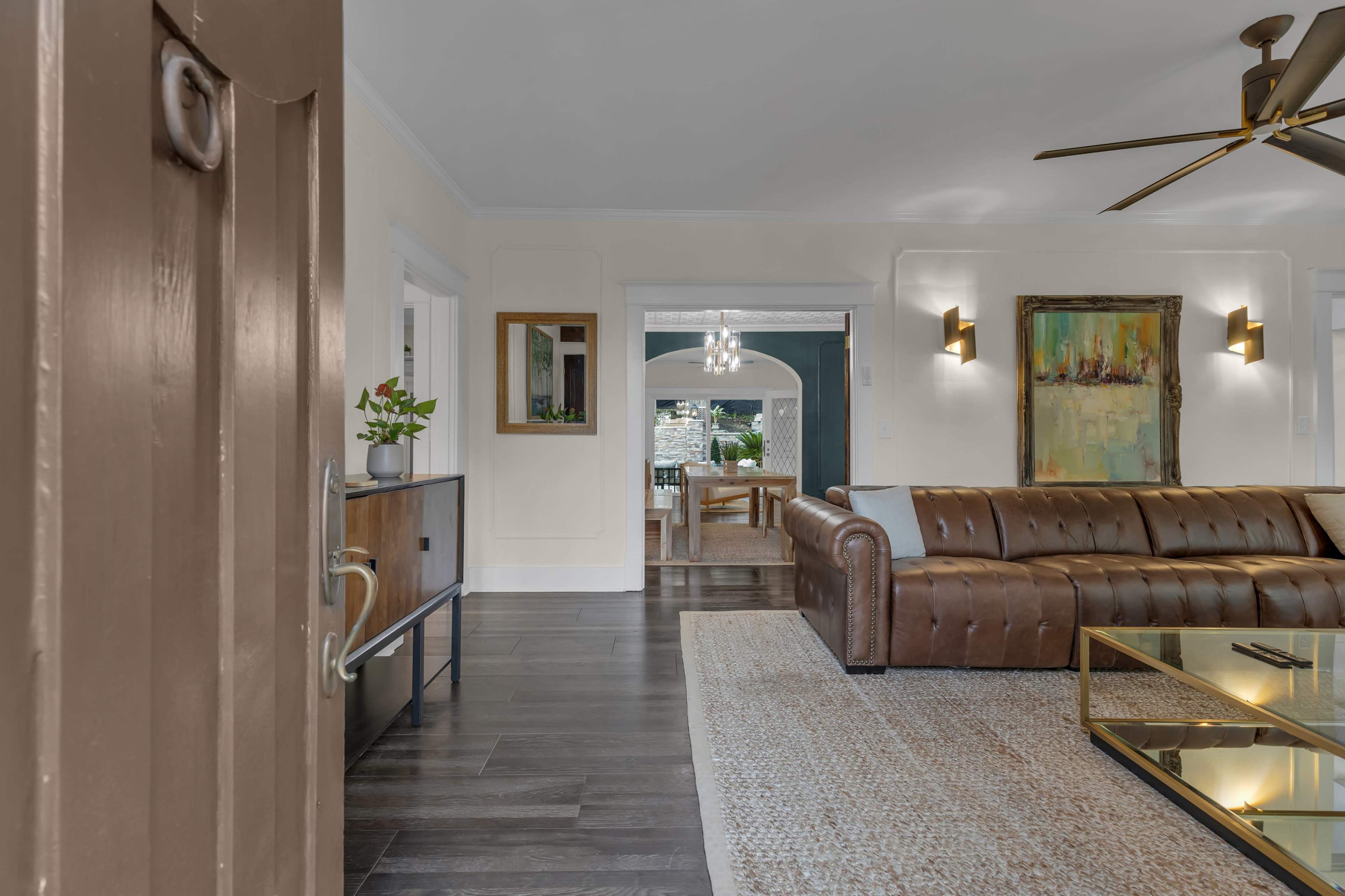 An interior living room with a brown leather sofa, a glass coffee table, and a view into a dining area with a chandelier and outdoor access.