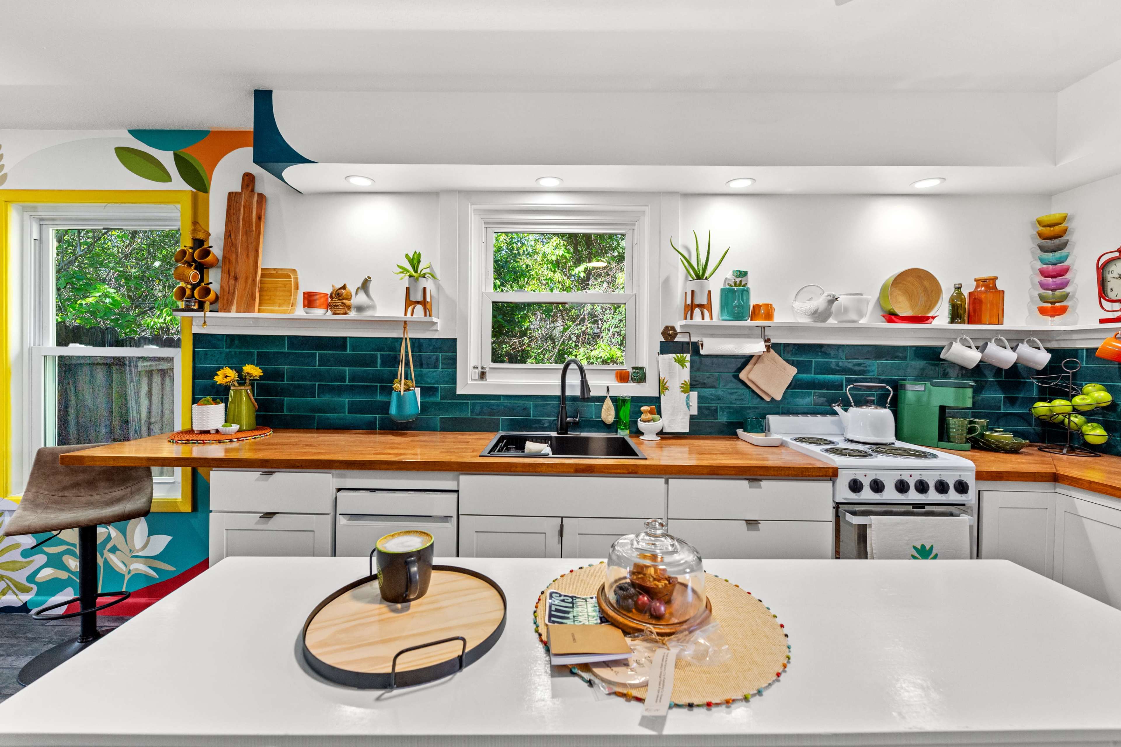 The kitchen features a mix of colorful wall decor, wooden accents, and modern appliances, with a central island displaying a tray and a glass container.