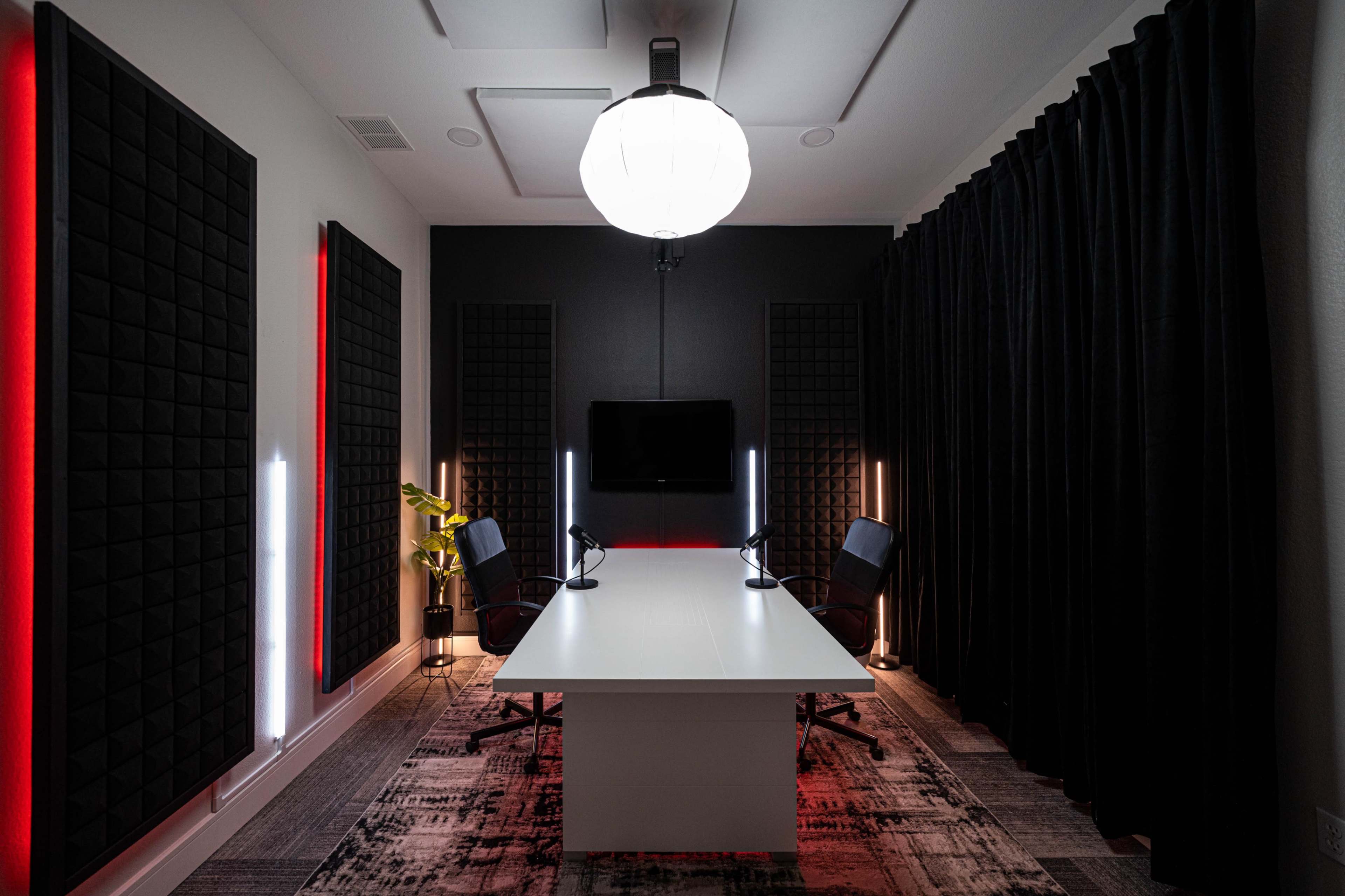 A modern conference room with two black chairs and a large white table, illuminated by a pendant light and accented with black soundproof panels and red LED lights.