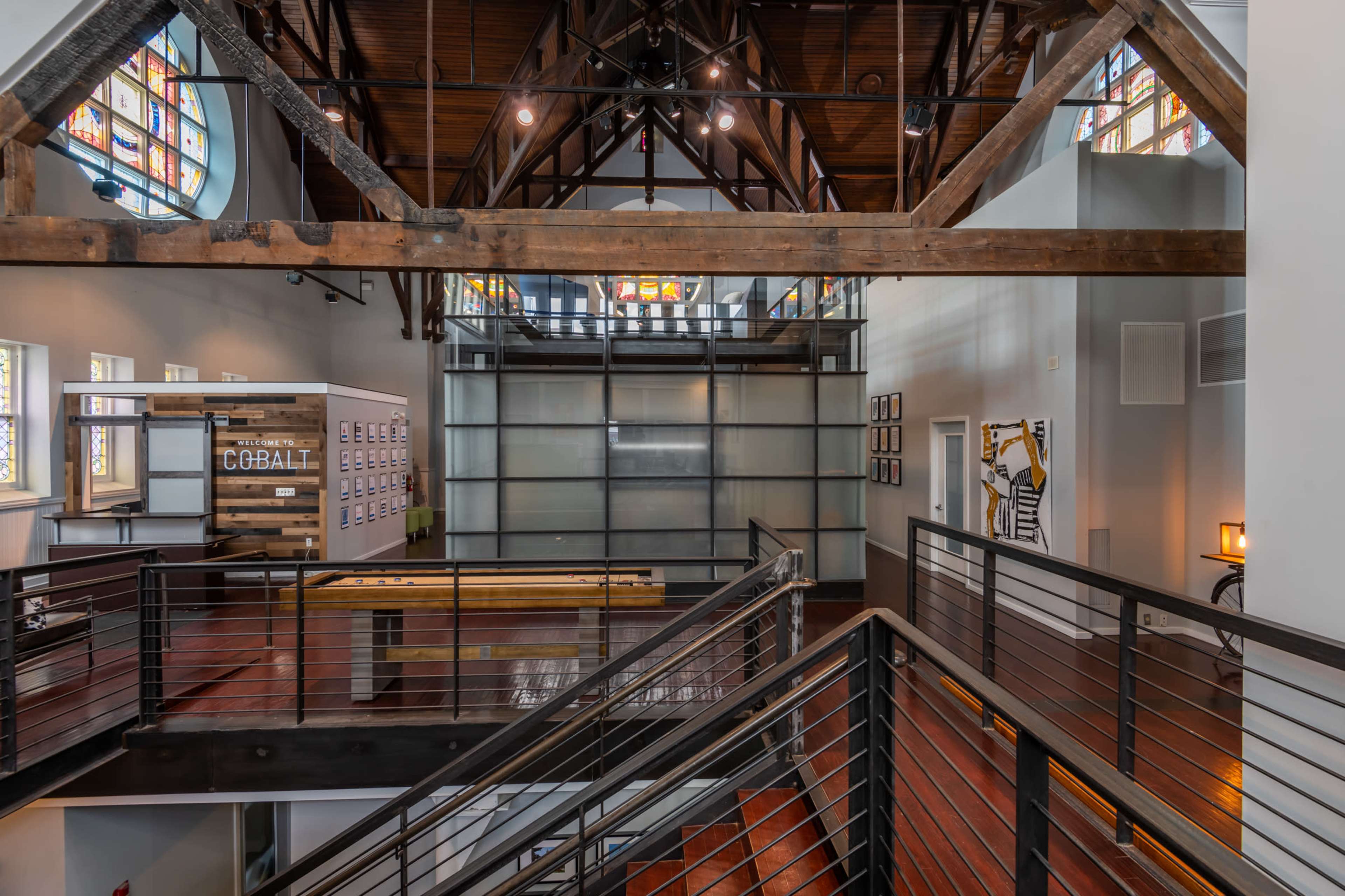The image shows the interior of a spacious, modern building with a high ceiling supported by wooden beams and large windows.