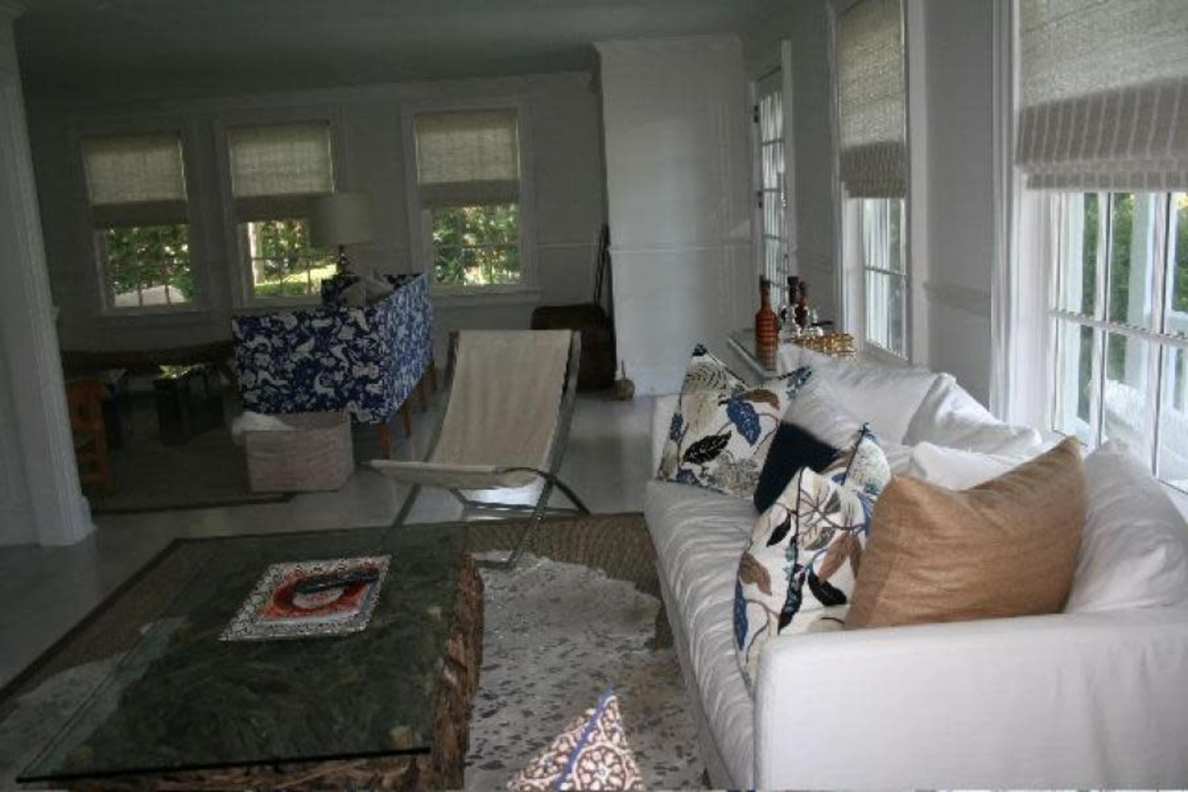 The image shows a living room with a white sofa, a glass coffee table, and large windows allowing natural light to enter the space.