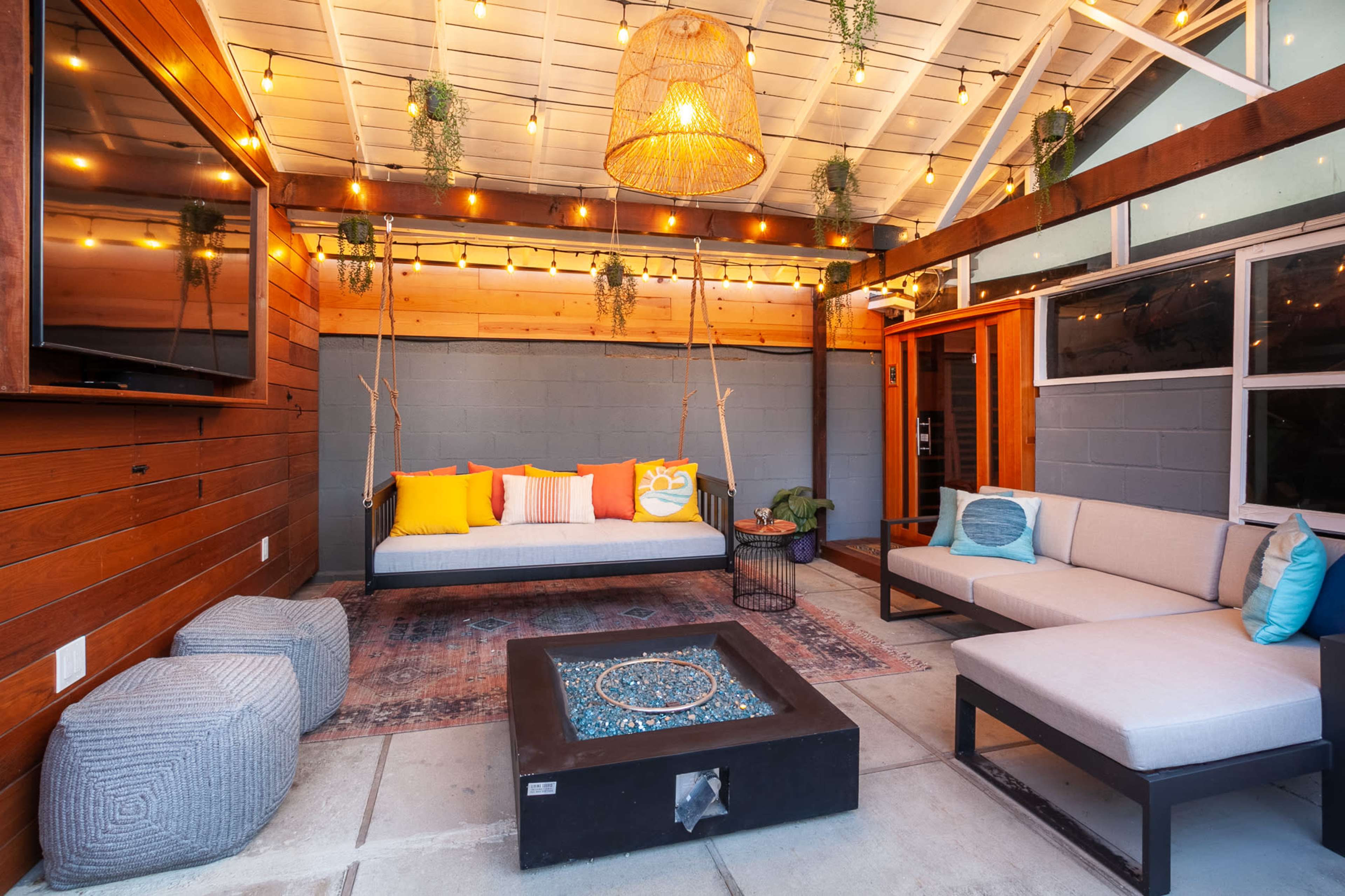 The image shows a cozy outdoor lounge area featuring a modern seating arrangement, a fire pit, and decorative string lights overhead.