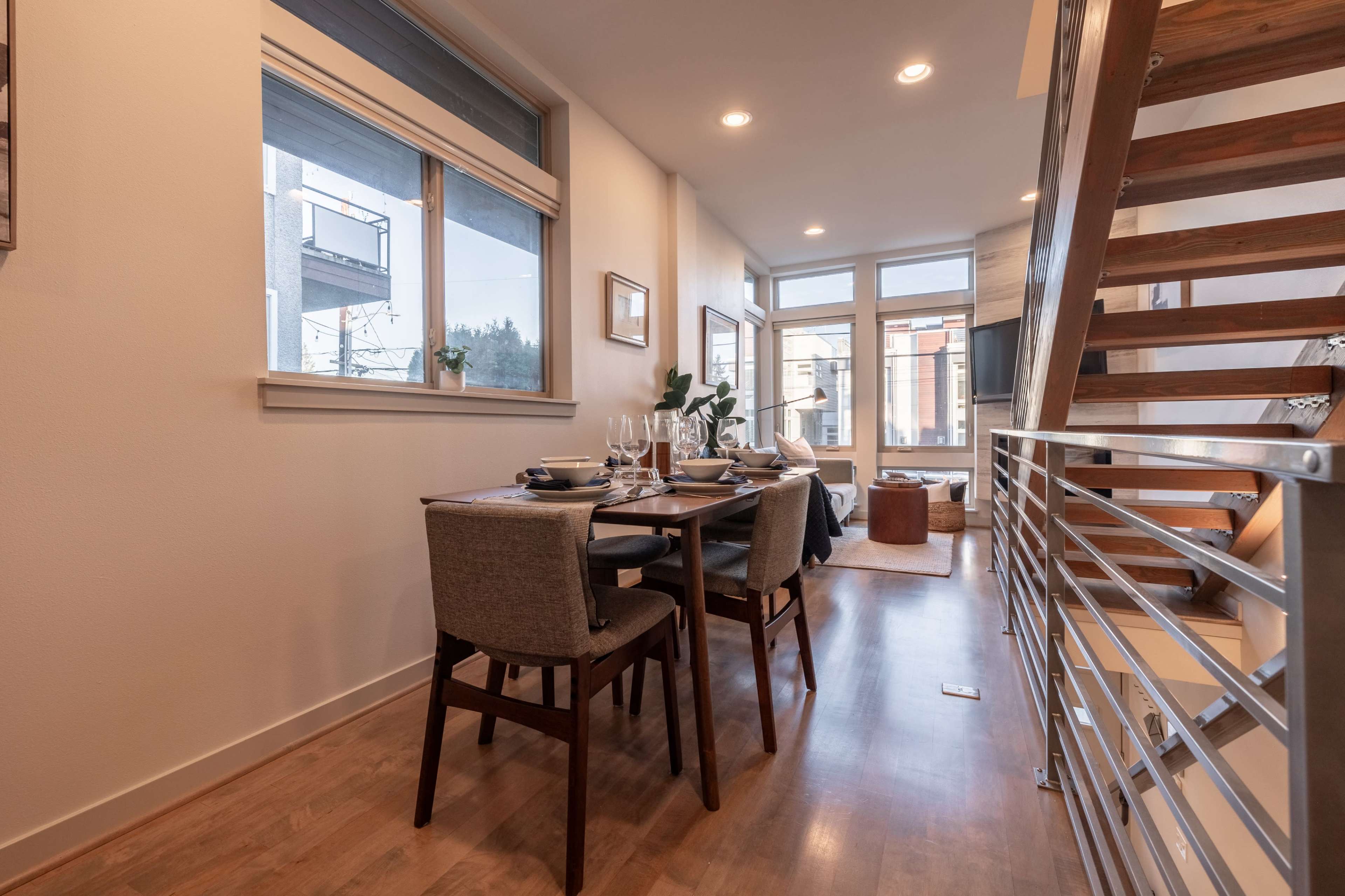 A dining area features a table set for four, with large windows and a view of a staircase leading to an upper level.