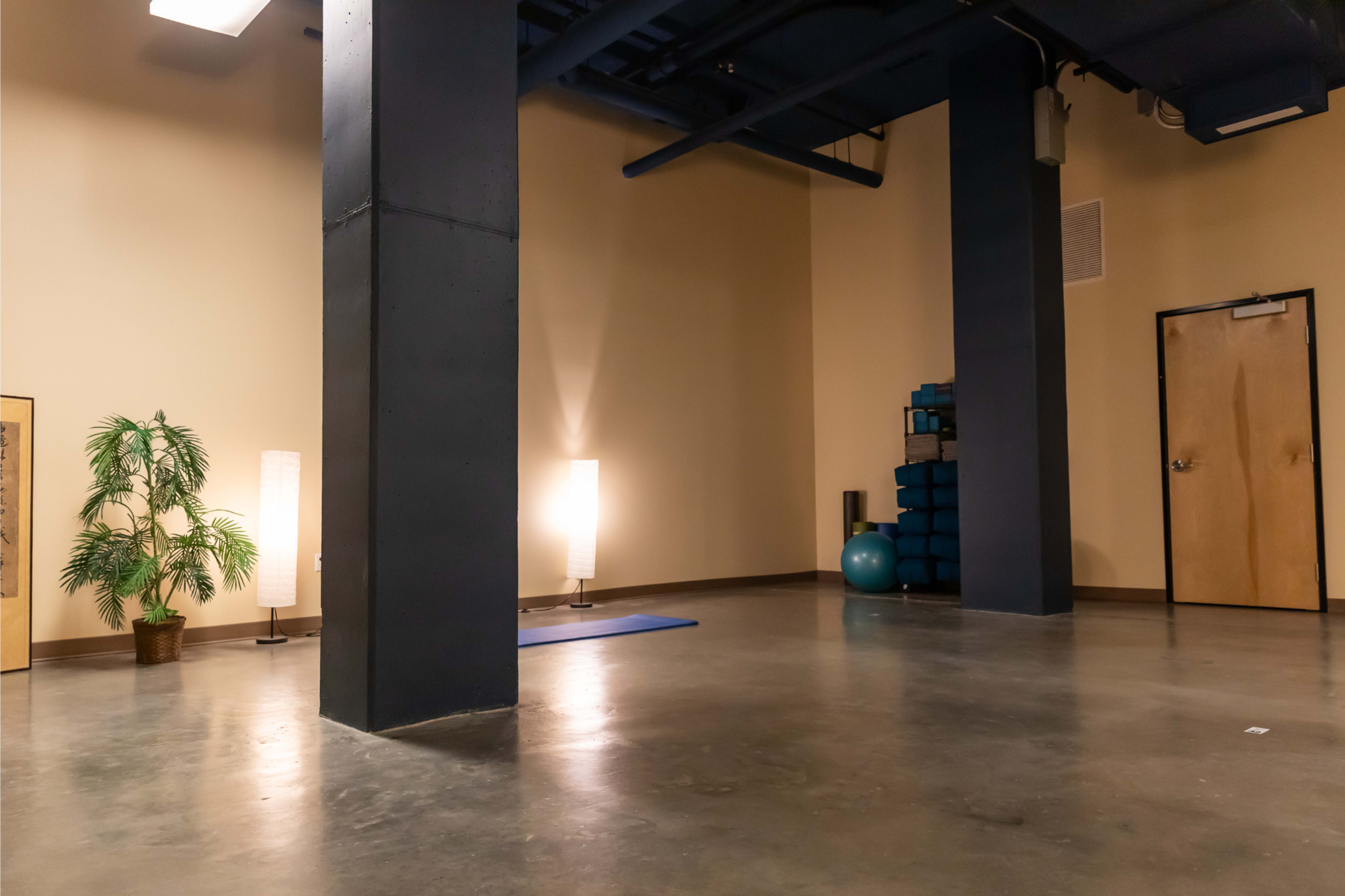 A spacious room with concrete flooring, featuring a large black column, yoga mats, a blue exercise ball, and soft lighting from floor lamps.