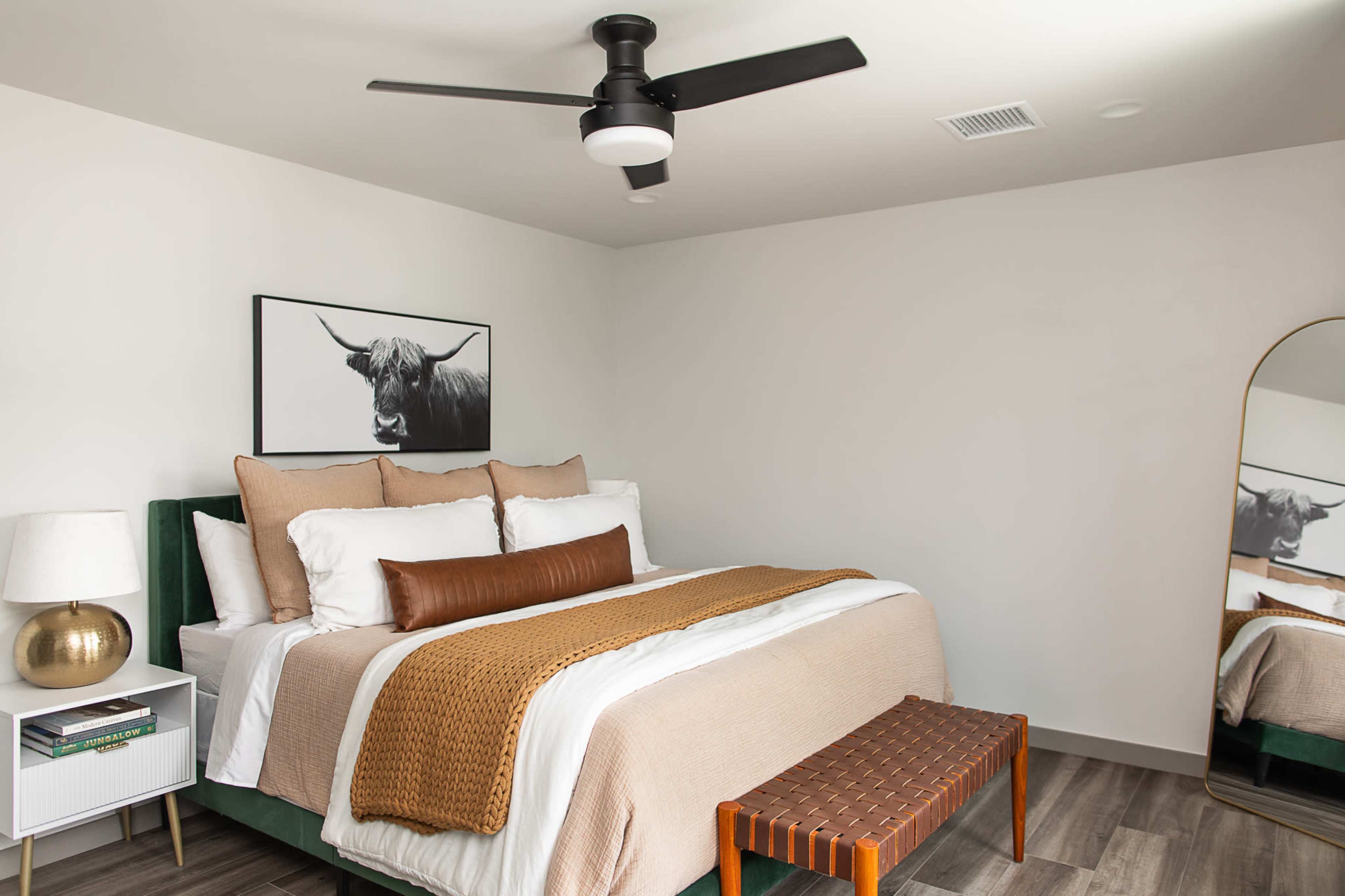 A modern bedroom featuring a queen-sized bed with white and beige bedding, a decorative brown lumbar pillow, a wooden bench at the foot of the bed, and a large mirror on one wall.
