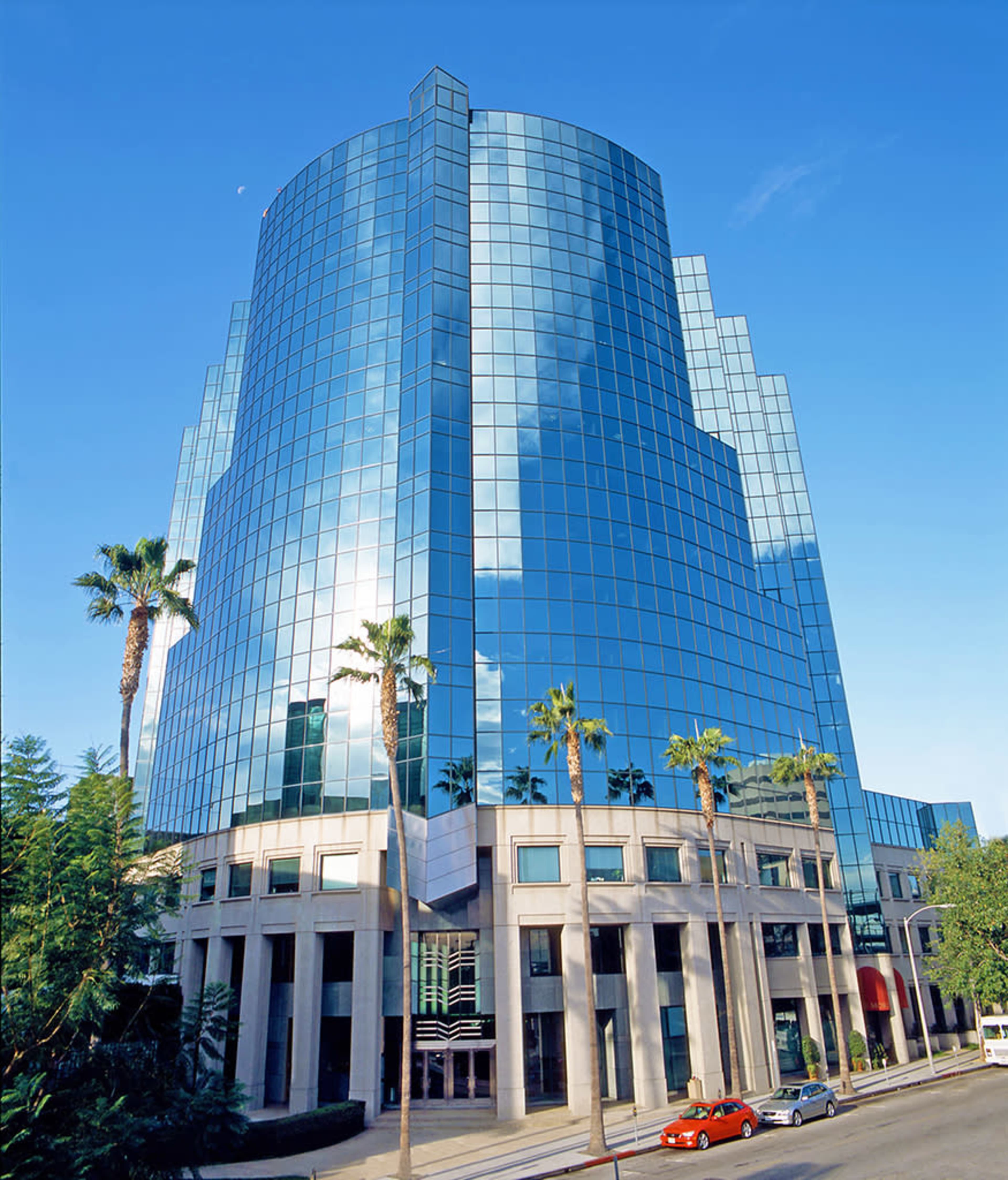 A modern, glass-covered office building with palm trees in the foreground and a clear blue sky.