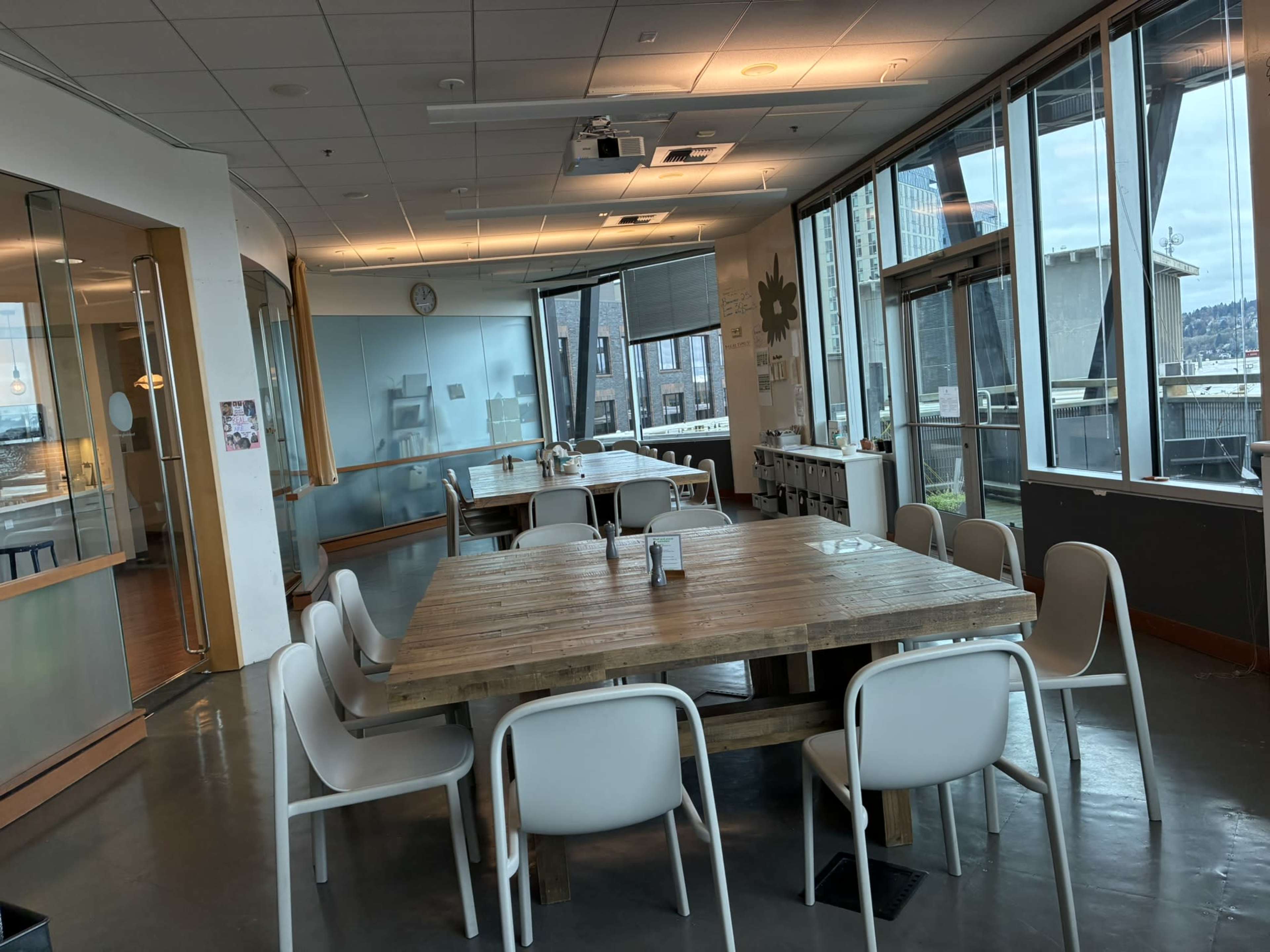 The image shows a spacious dining area with a large wooden table surrounded by white chairs, featuring large windows that provide a view of the outside.