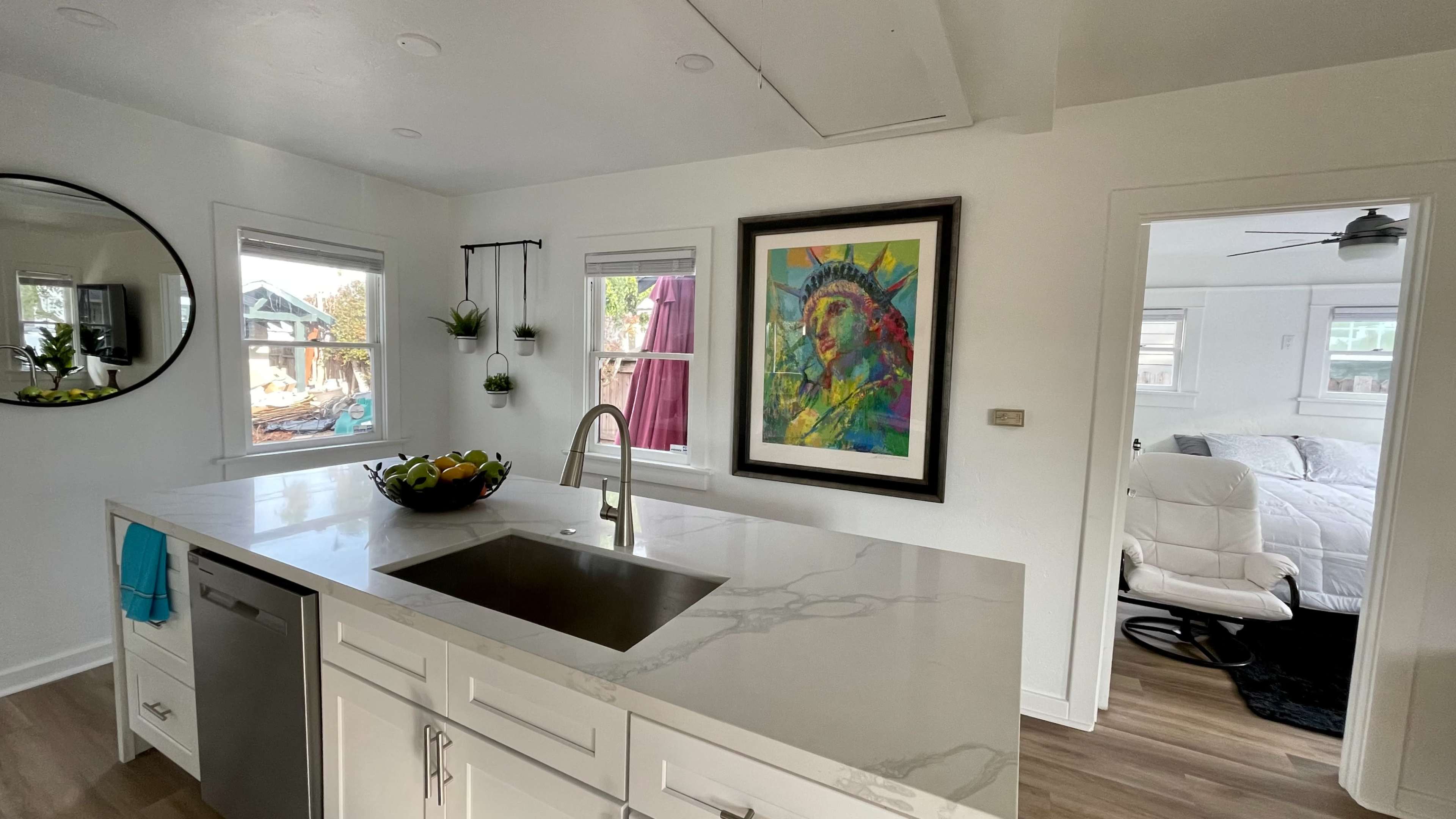A modern kitchen with a marble countertop, a sink, and a framed artwork of the Statue of Liberty on the wall, adjacent to a living area with a white chair and a bed.