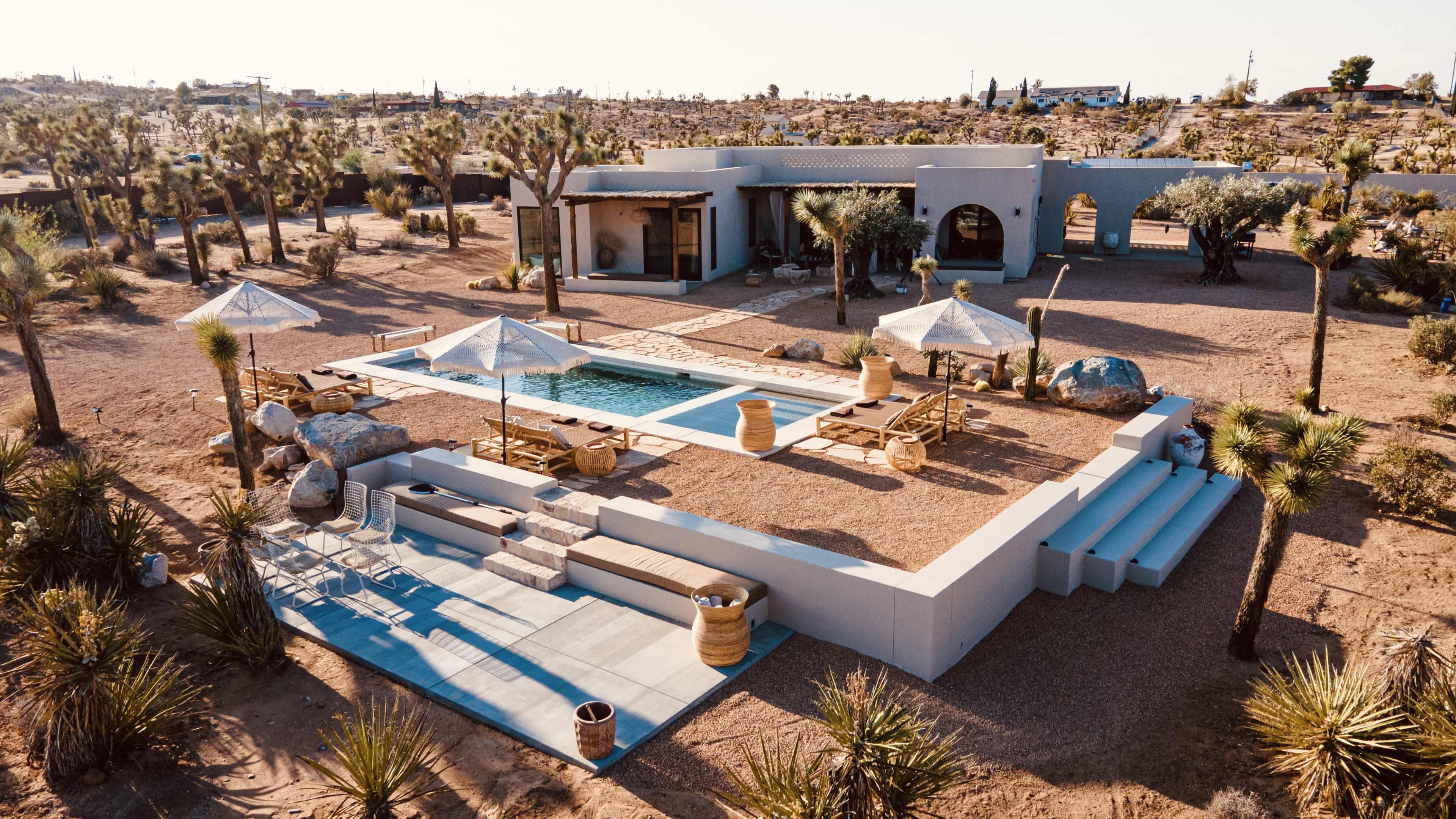 A modern desert house with a pool, surrounded by landscaping featuring Joshua trees and outdoor seating areas.