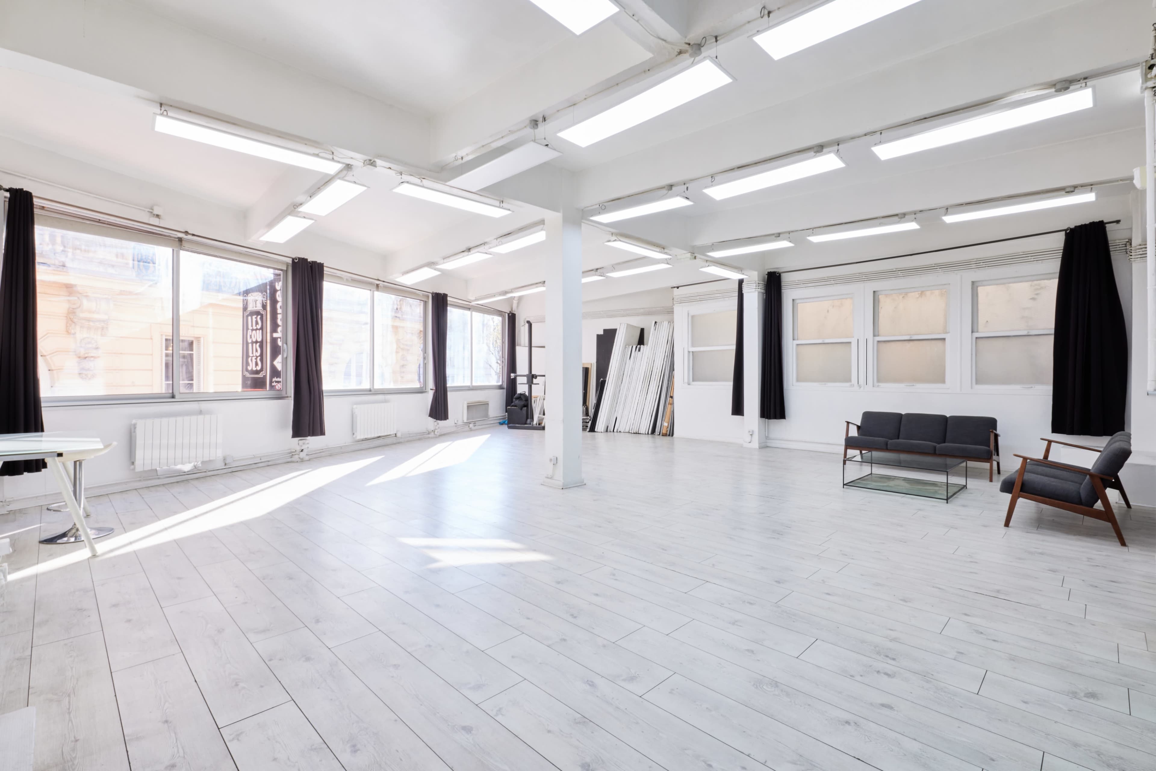The image shows a spacious, well-lit studio with large windows, white walls, and a hardwood floor, featuring minimal furniture including a couch and a chair.