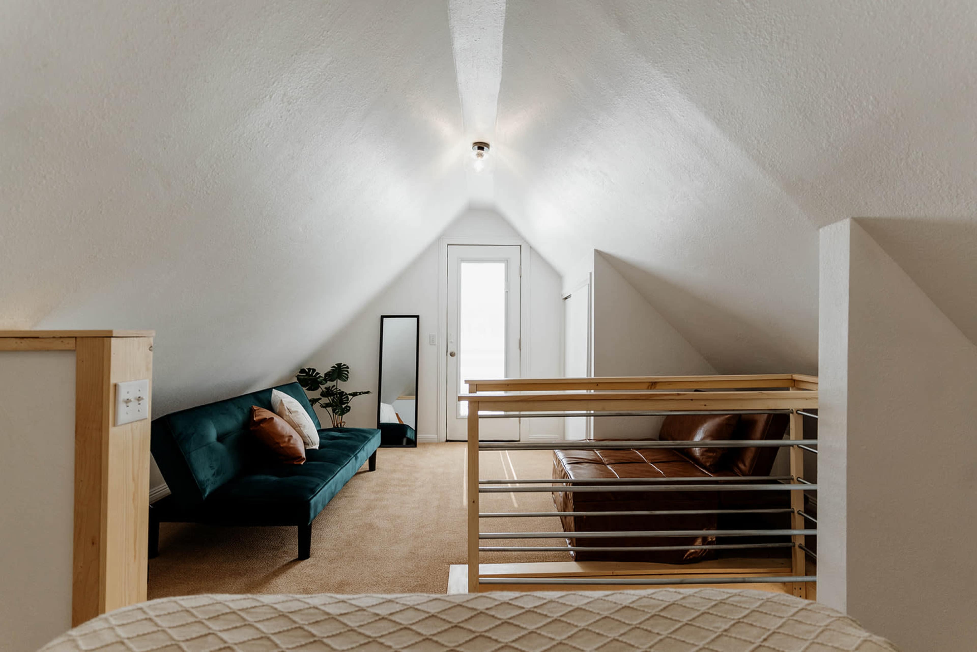 An airy attic space furnished with a teal couch and a brown leather sofa, featuring a door and a mirror against the white walls.