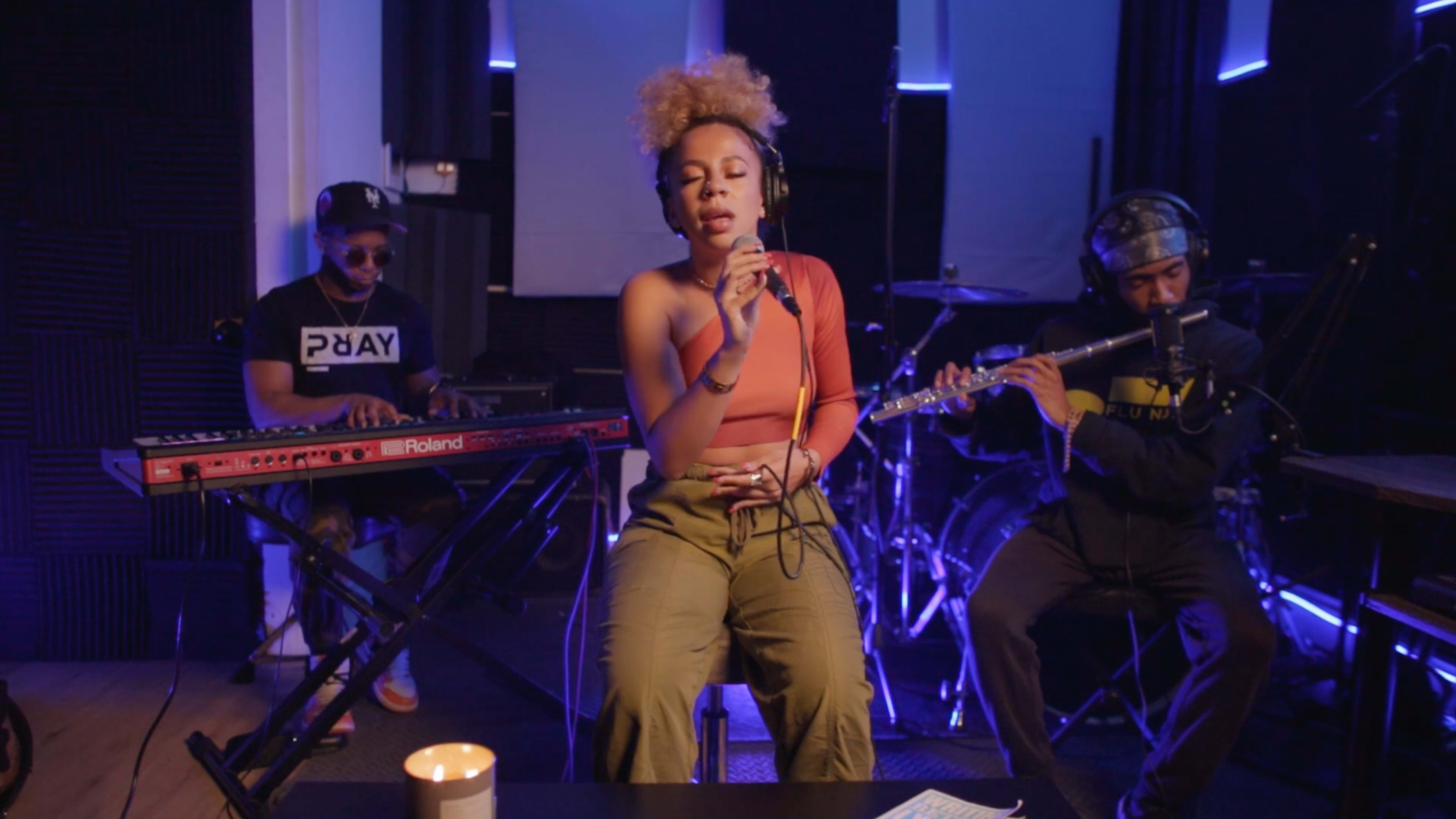 A woman sings into a microphone while seated in a studio, accompanied by a man playing a keyboard and another playing a flute.
