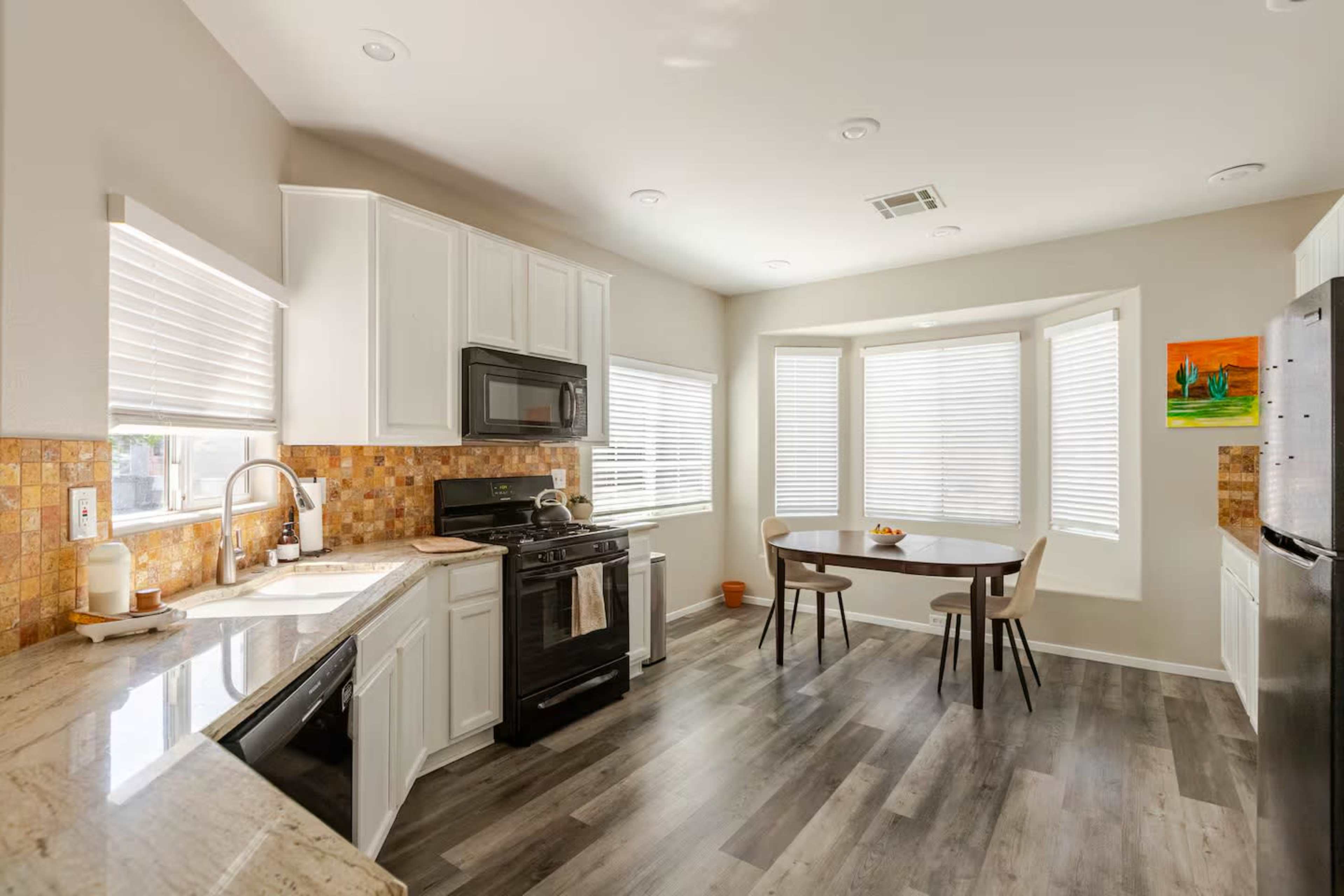 The image shows a modern kitchen with white cabinets, stainless steel appliances, and a dining area featuring a round table near large windows.