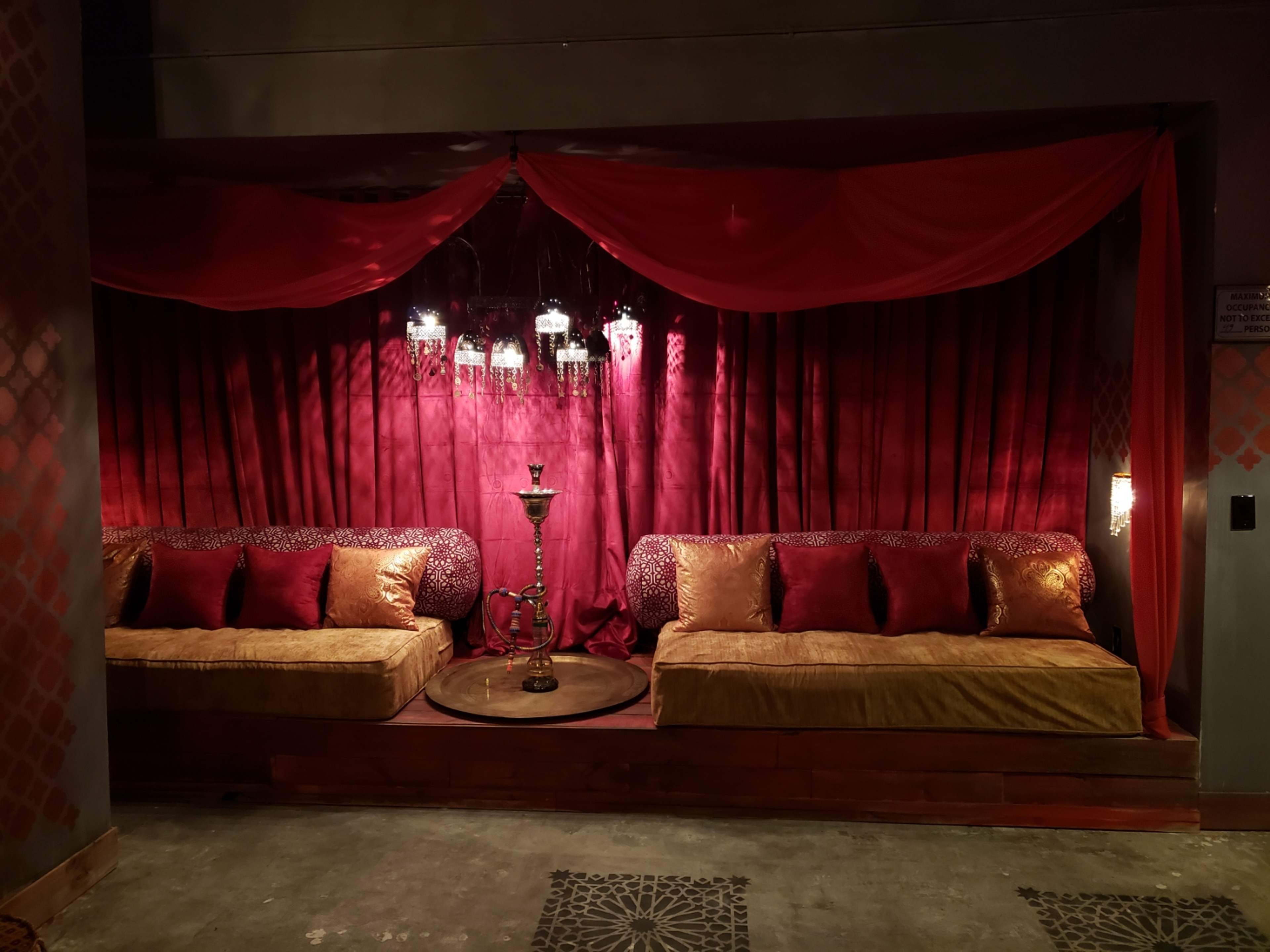 A seating area with two plush couches, adorned with red cushions, set against a burgundy draped backdrop and illuminated by hanging light fixtures.