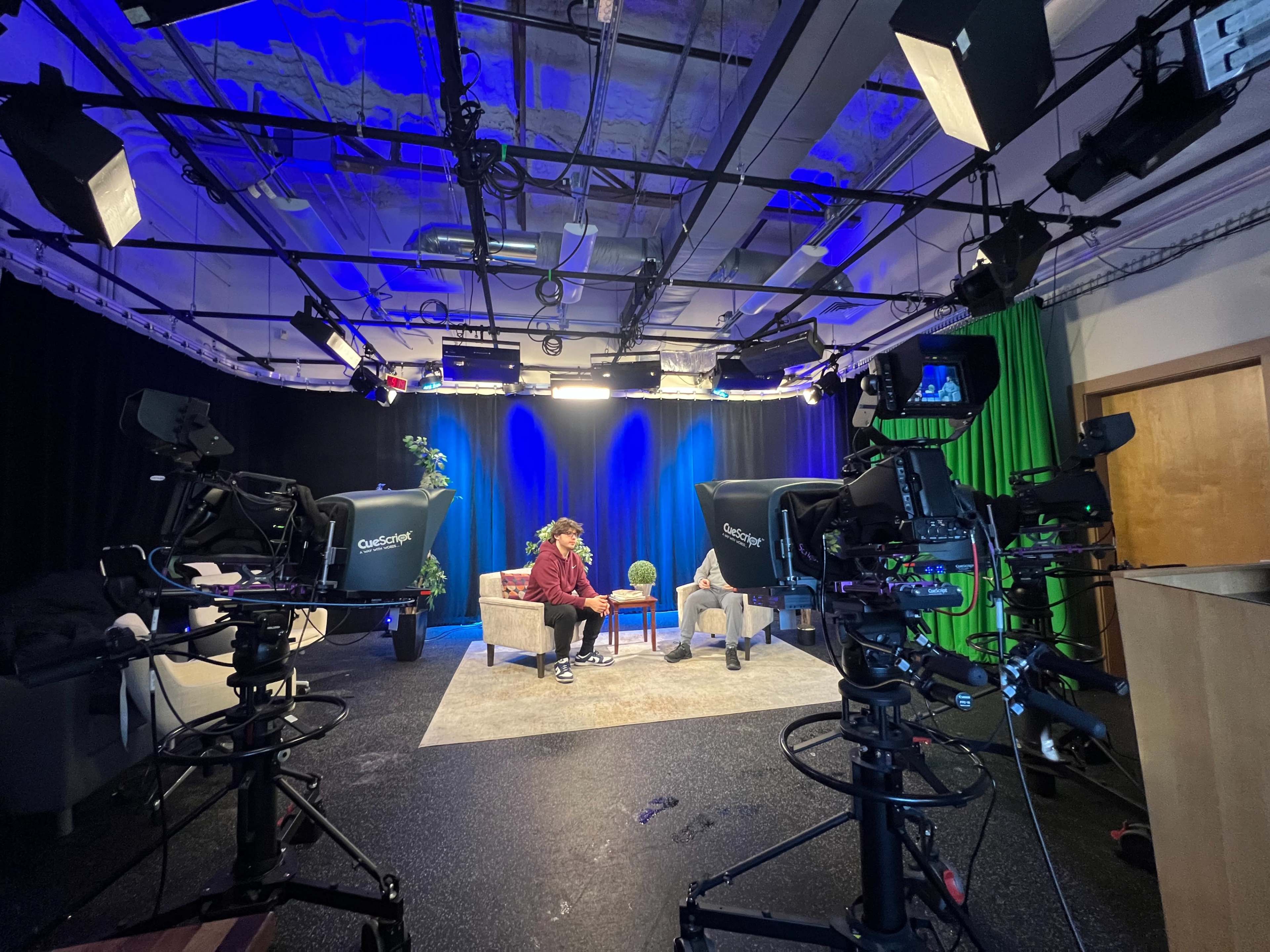 A television studio is set up with two cameras focused on a person sitting on a couch, surrounded by studio lights and a blue curtain backdrop.