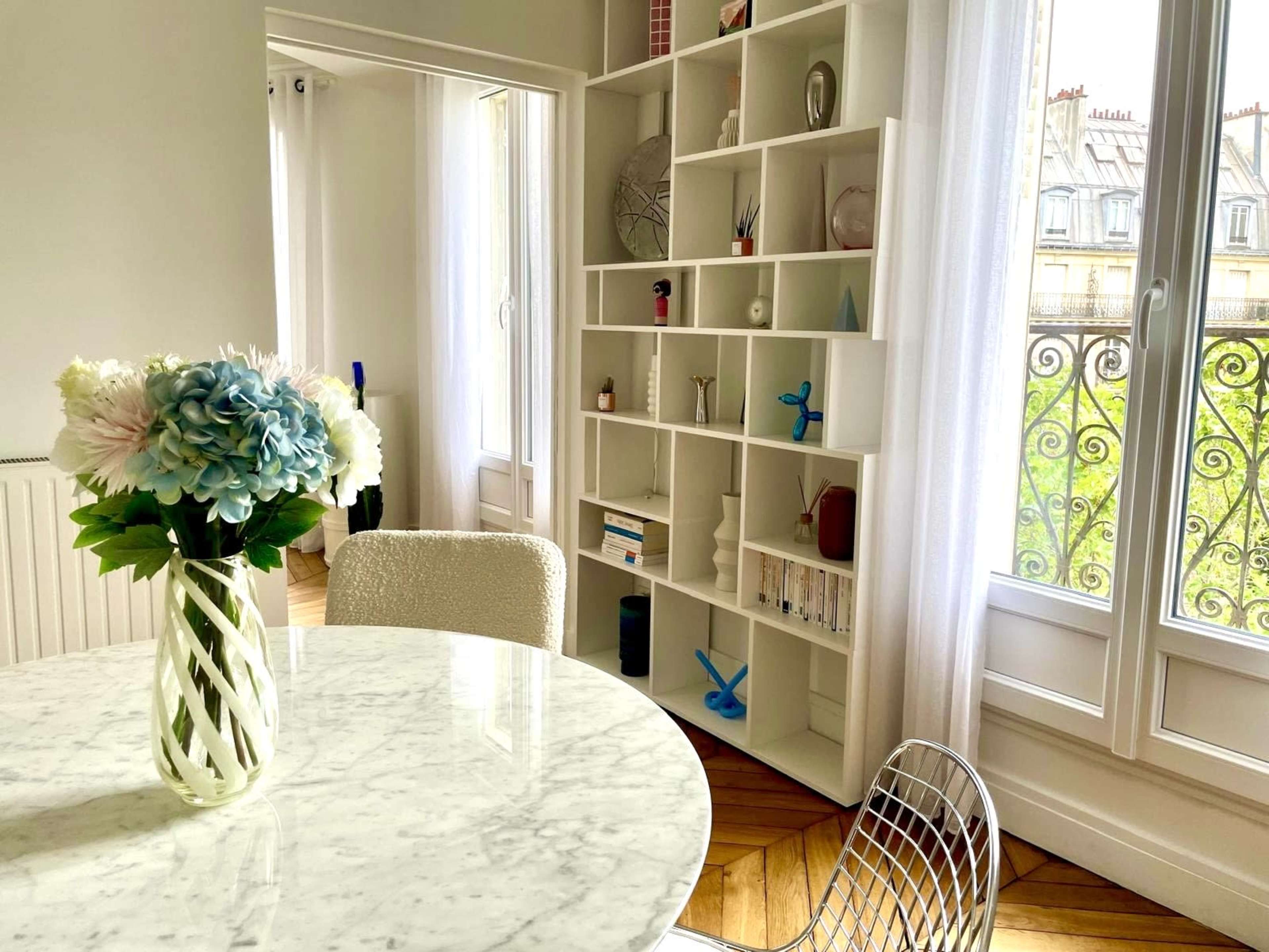 A round marble dining table is set in a bright room with a tall bookshelf against the wall and a vase of flowers on the table.
