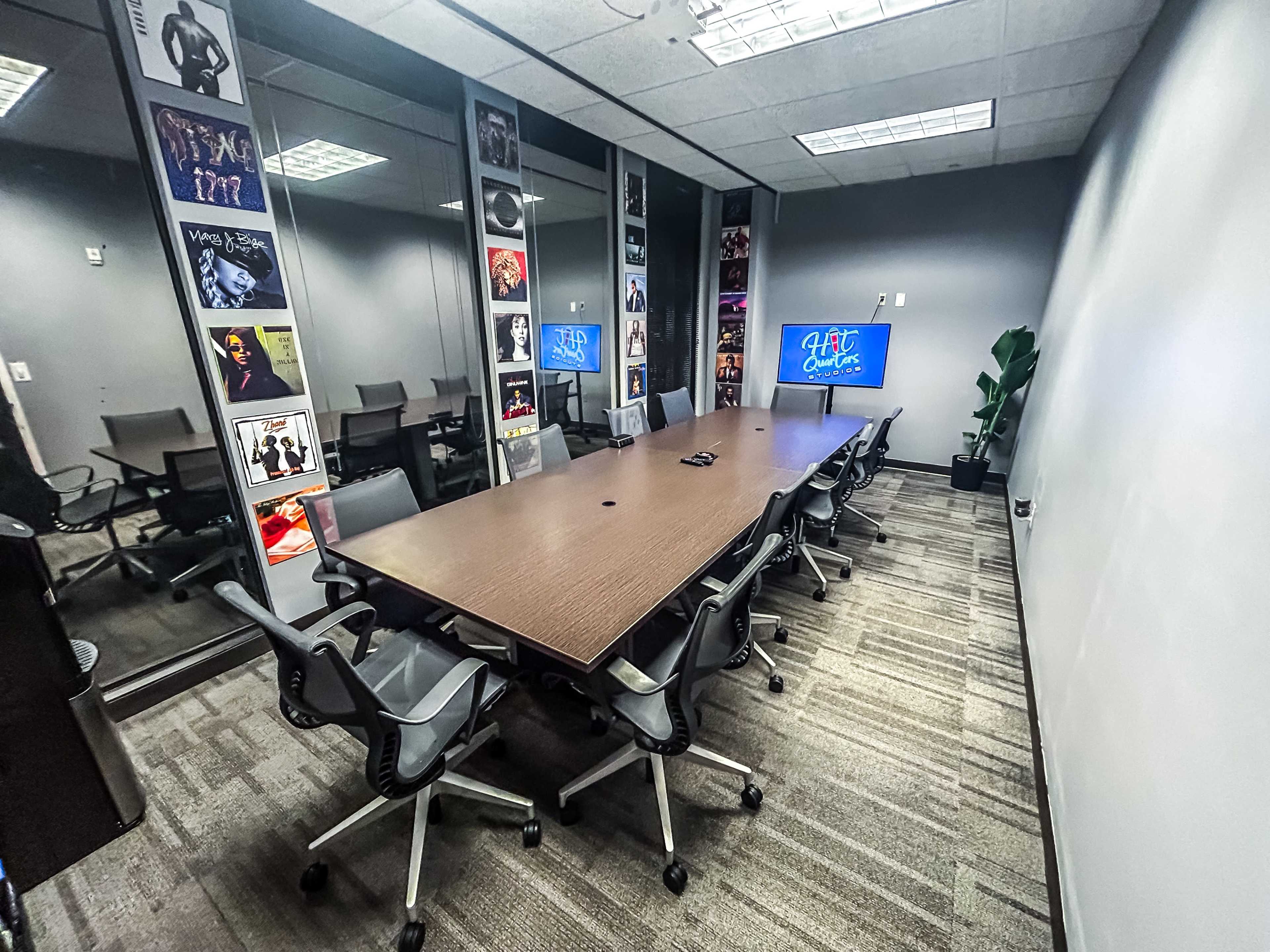 A modern conference room features a large wooden table surrounded by ergonomic chairs, with glass walls displaying framed posters and a television screen.