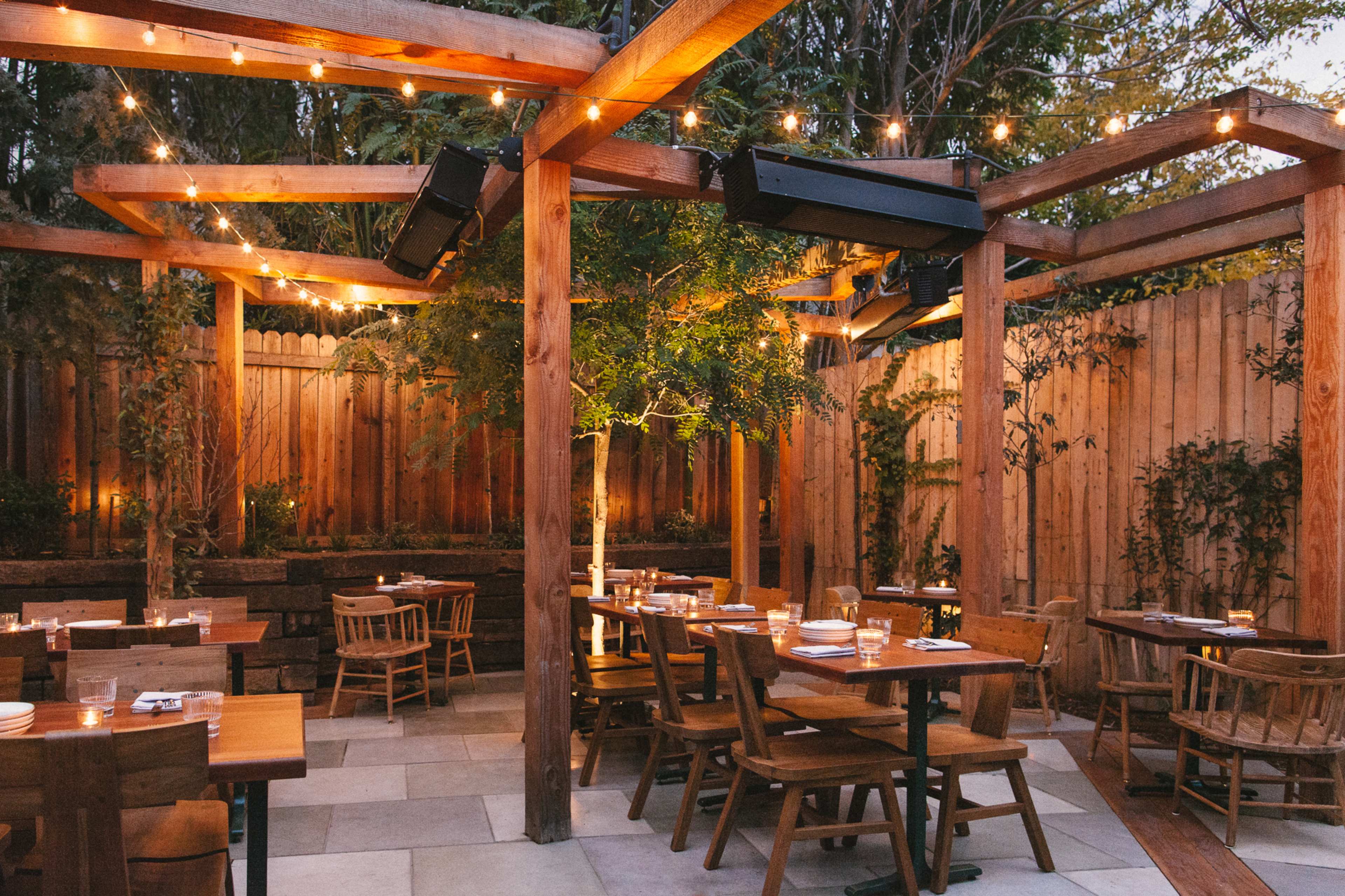 The image shows an outdoor restaurant patio with wooden tables and chairs arranged under string lights and surrounded by greenery.