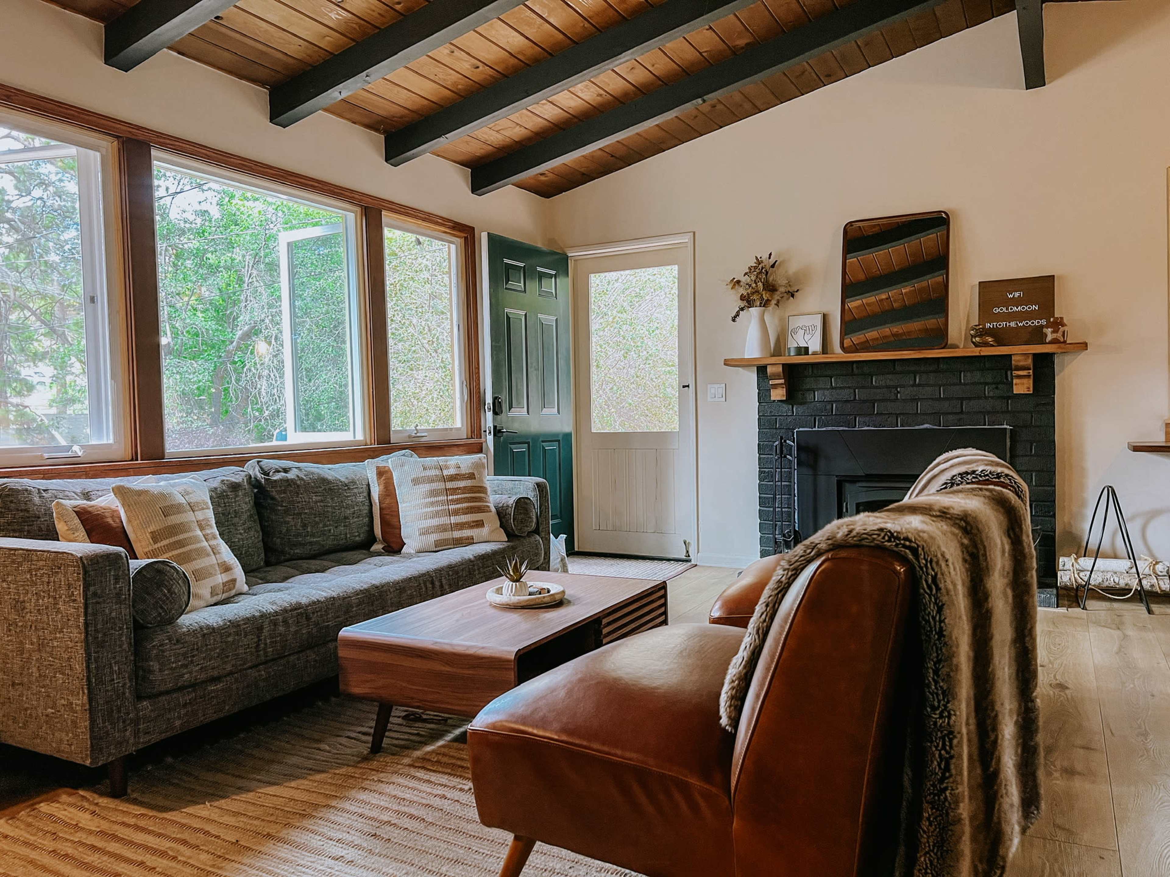 The image shows a living room with large windows, a gray sofa, brown armchairs, a wooden coffee table, and a fireplace against a black brick wall.