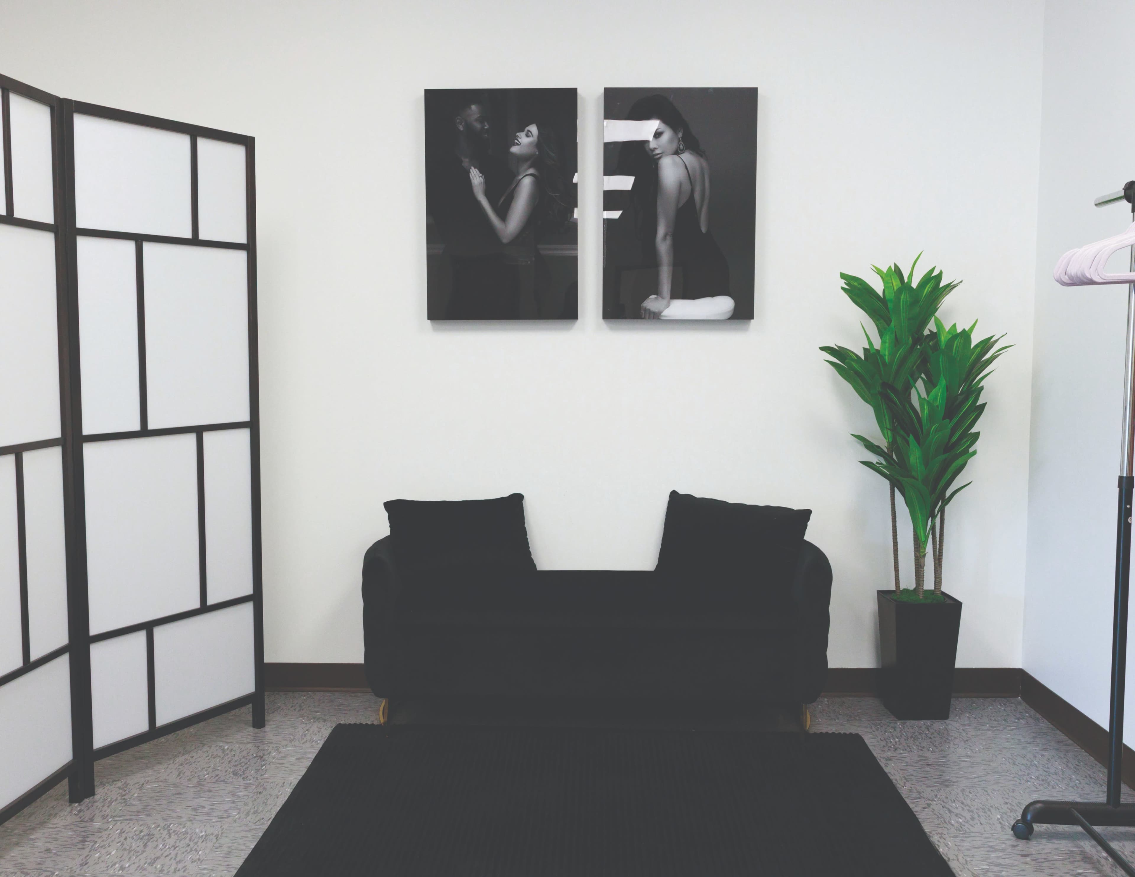 The image shows a minimalist interior space with a black sofa, two framed black and white portraits on the wall, a potted plant, and a folding screen.