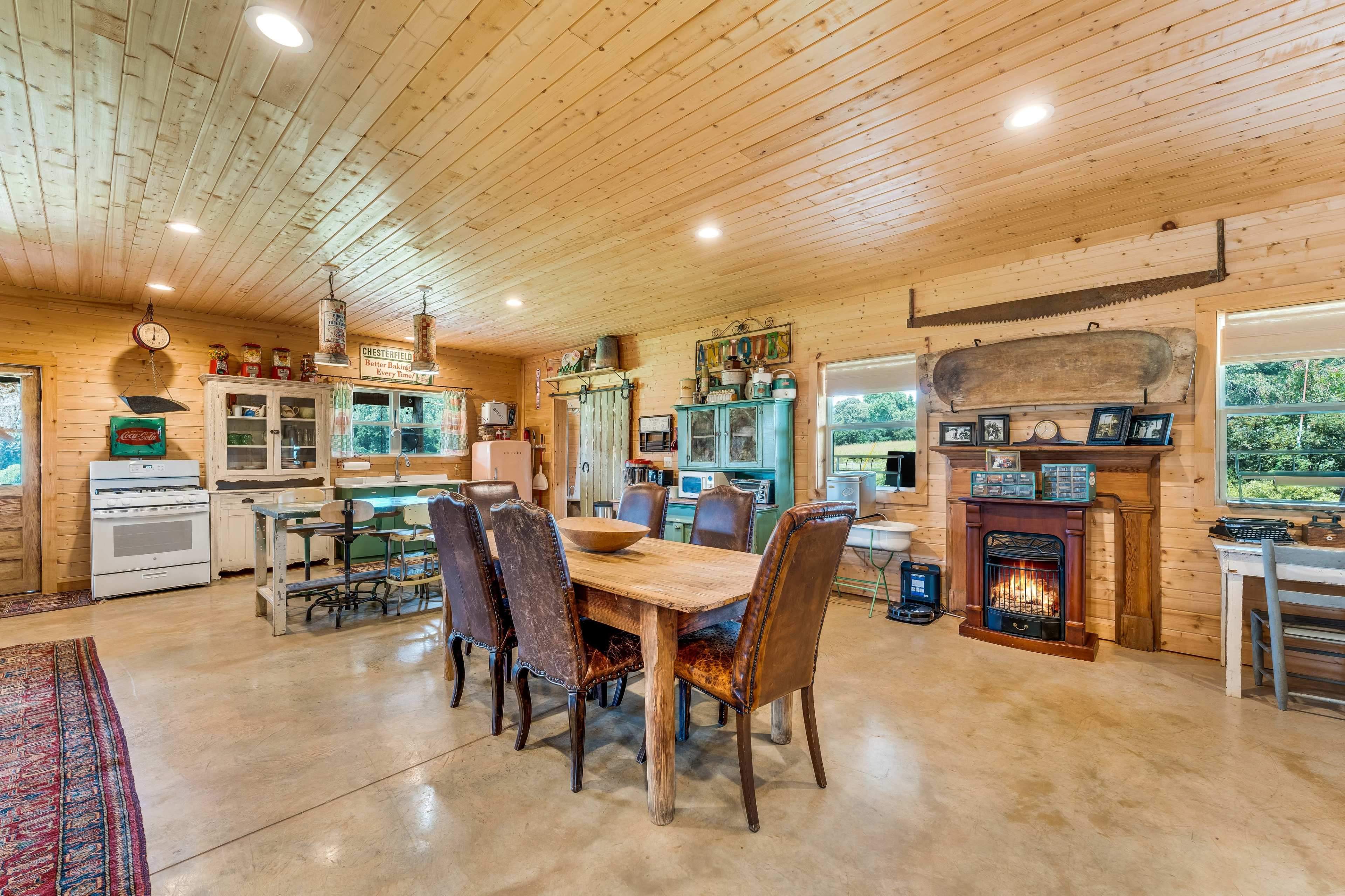 The image shows a spacious, rustic kitchen and dining area with wooden walls, a large dining table, and a cozy fireplace.