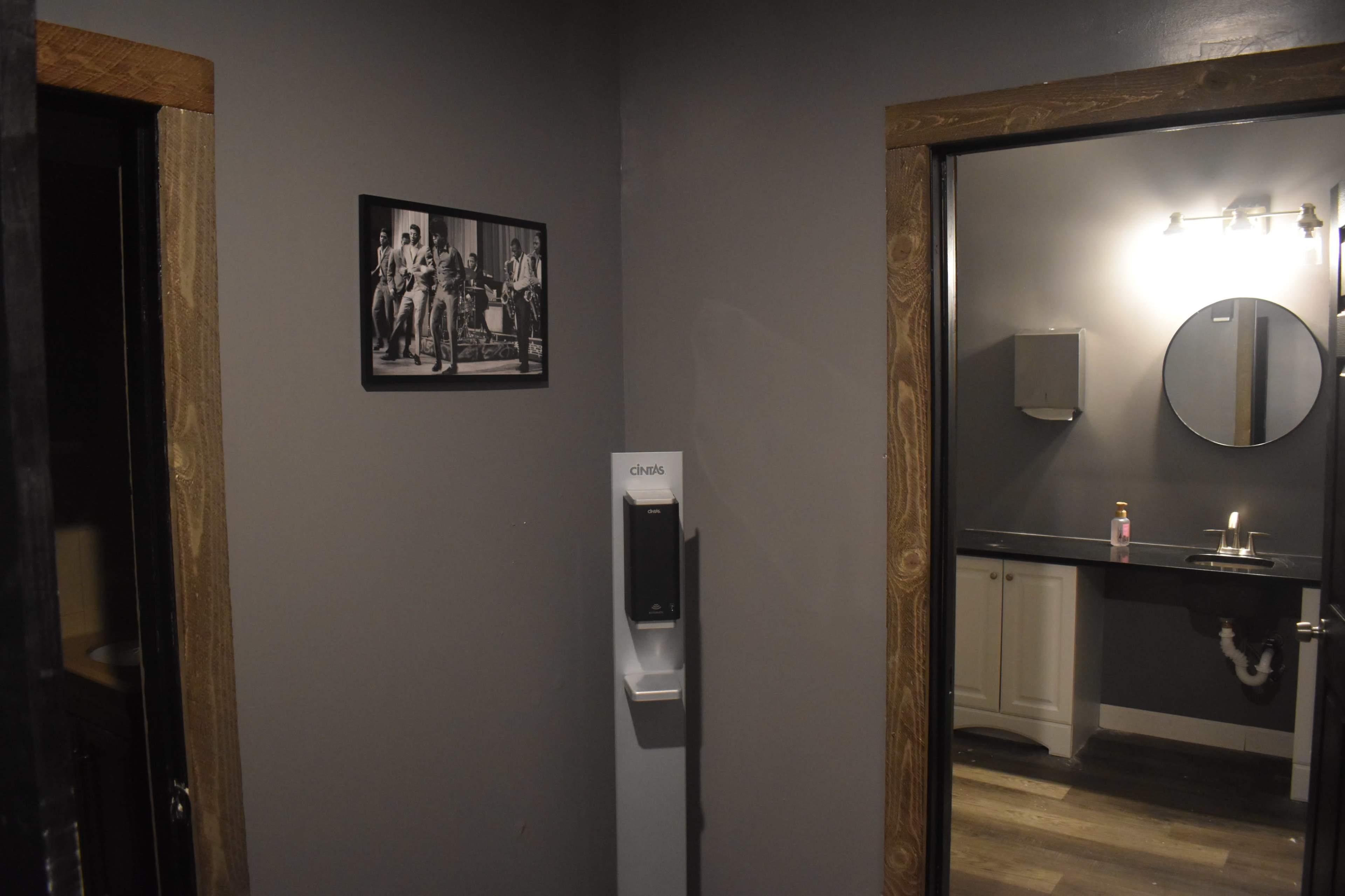 A dimly lit restroom area with a gray wall, featuring a framed black-and-white photograph, a soap dispenser, and a mirror above a sink.