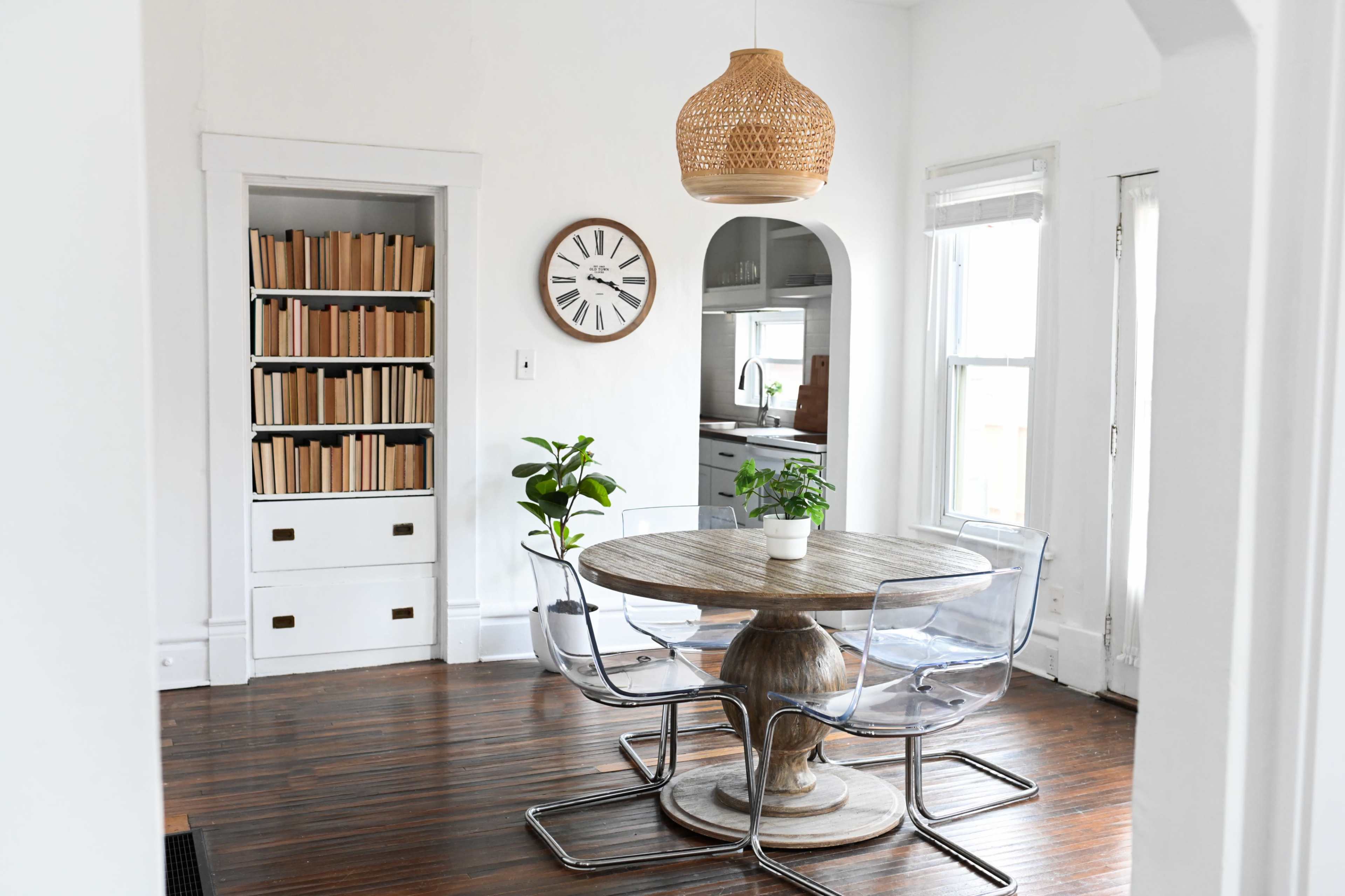 A bright dining area with a round wooden table surrounded by transparent chairs, a clock on the wall, and a bookshelf filled with books.
