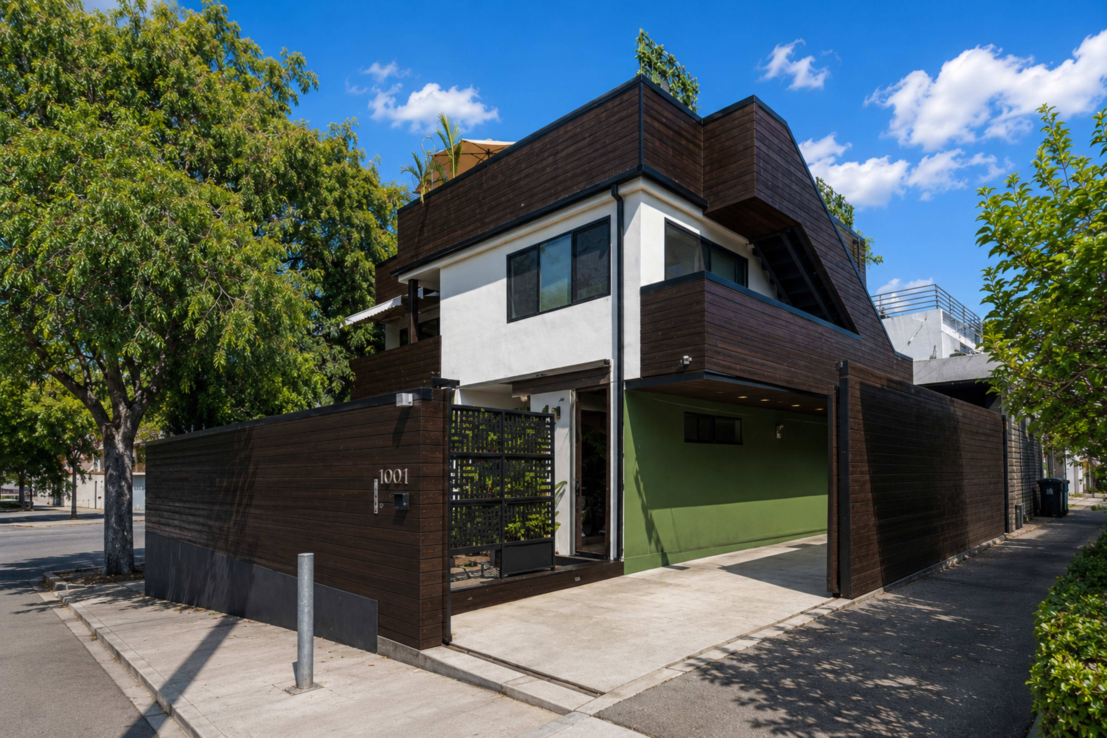 The image shows a modern two-story house with a dark wooden exterior, a green accent wall, and a gated entrance, situated on a tree-lined street.