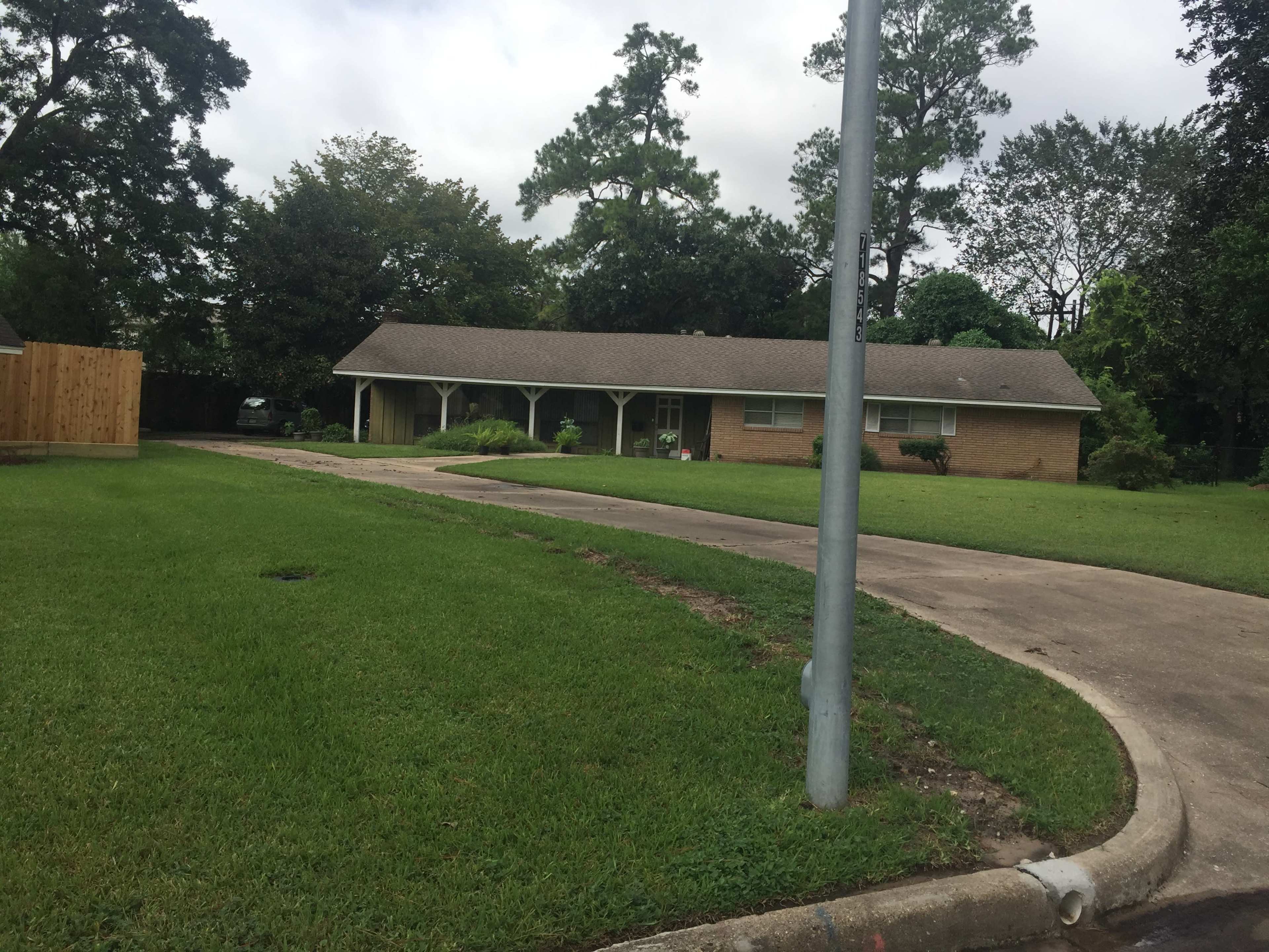 A single-story brick house with a front porch set on a grassy lawn, bordered by trees and a paved driveway.
