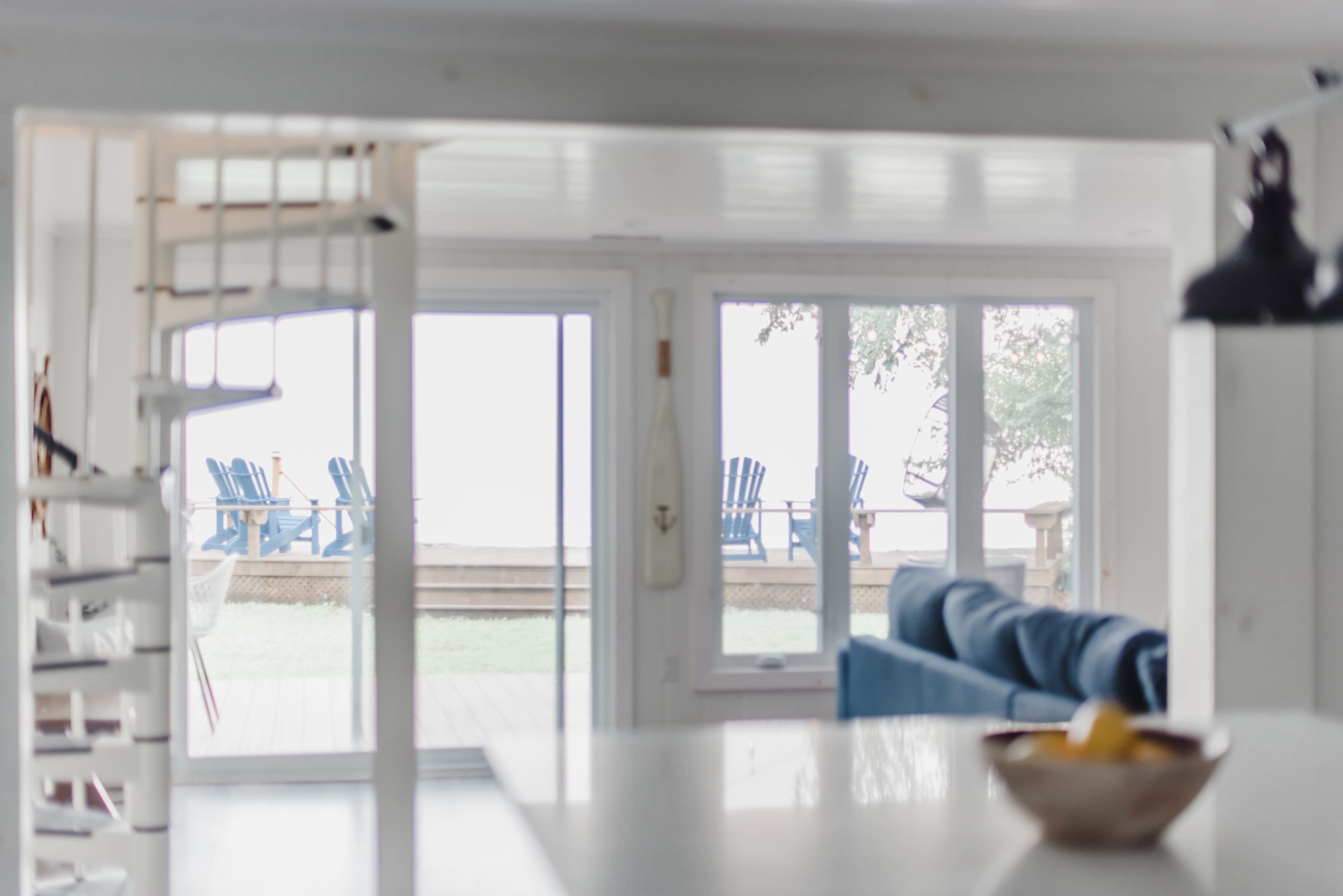 A spiral staircase leads to a bright room with large windows overlooking a deck featuring several blue chairs.