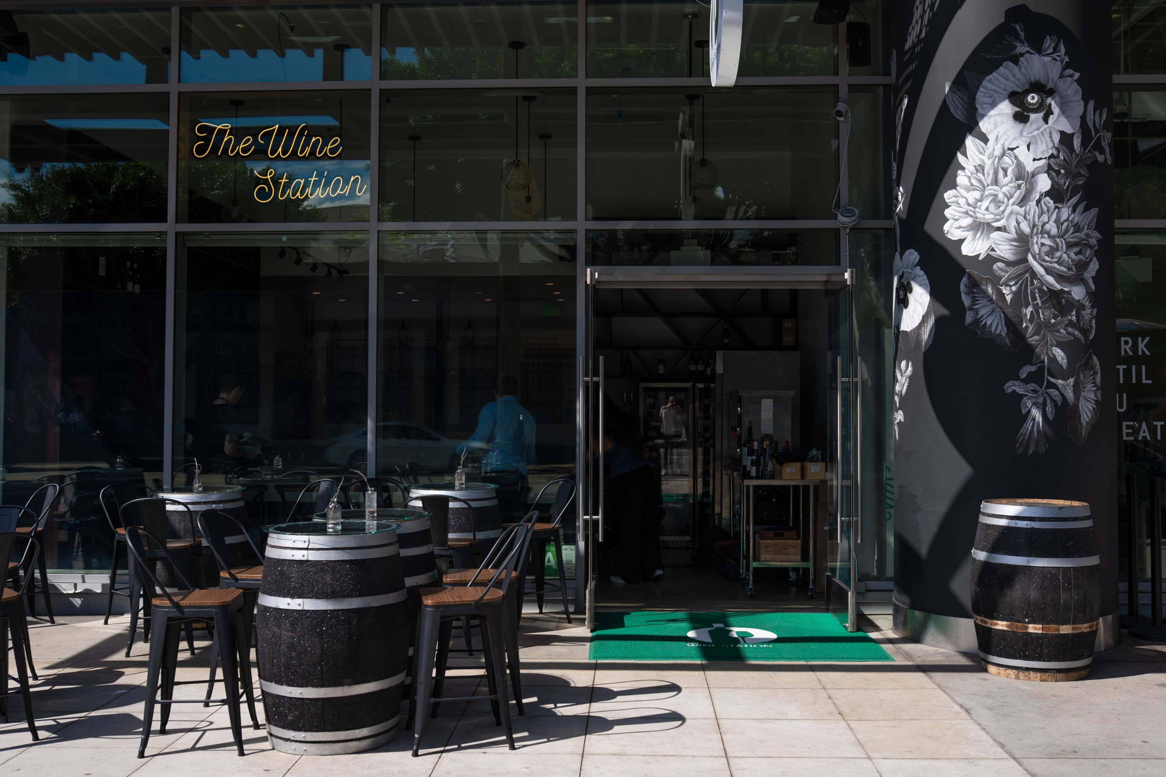 The image shows the exterior of a wine bar called "The Wine Station," featuring a large glass entrance, outdoor seating with black chairs, and decorative barrels.