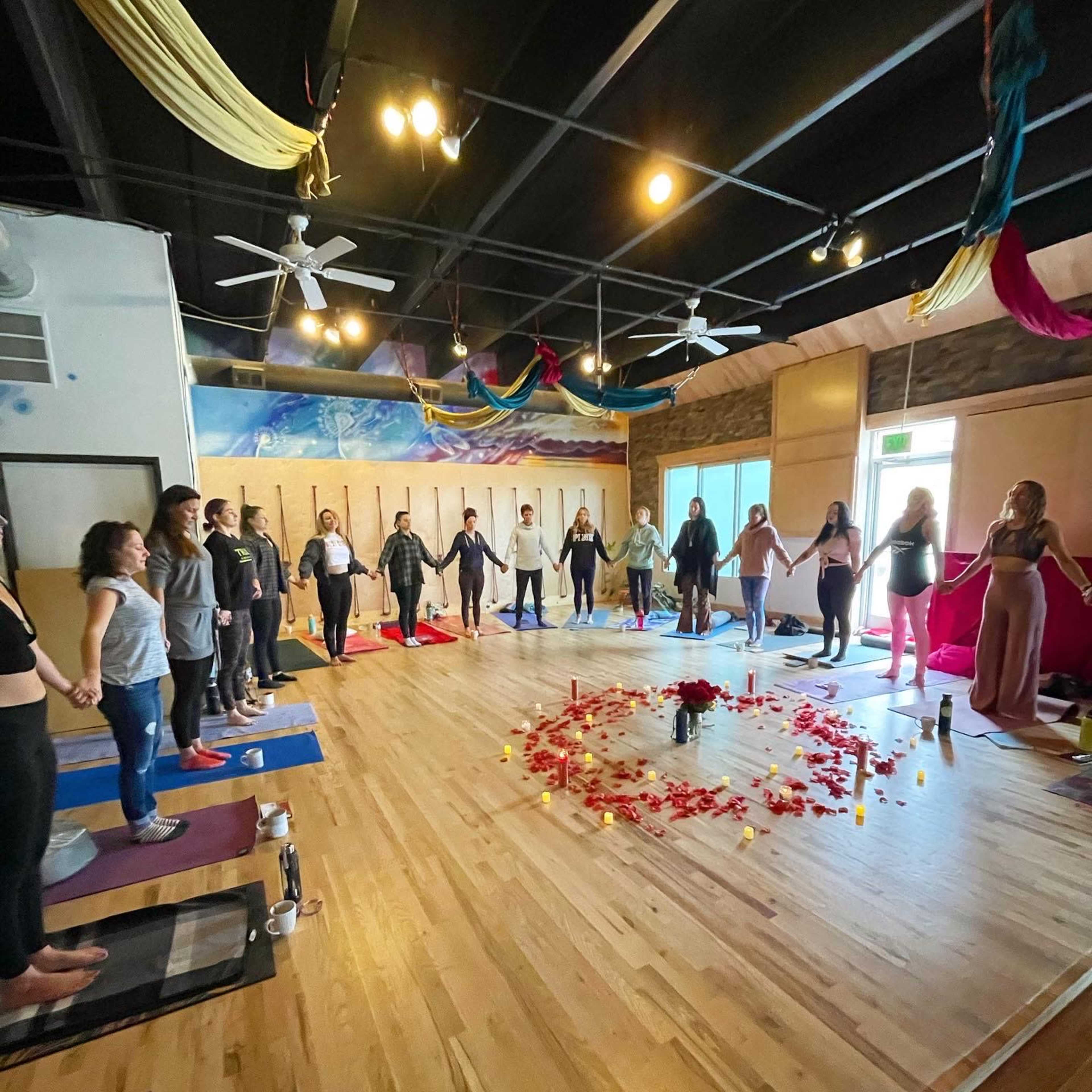 A group of people stands in a circle, holding hands around a flower and candle arrangement on the wooden floor of a spacious room with colorful decorations hanging from the ceiling.
