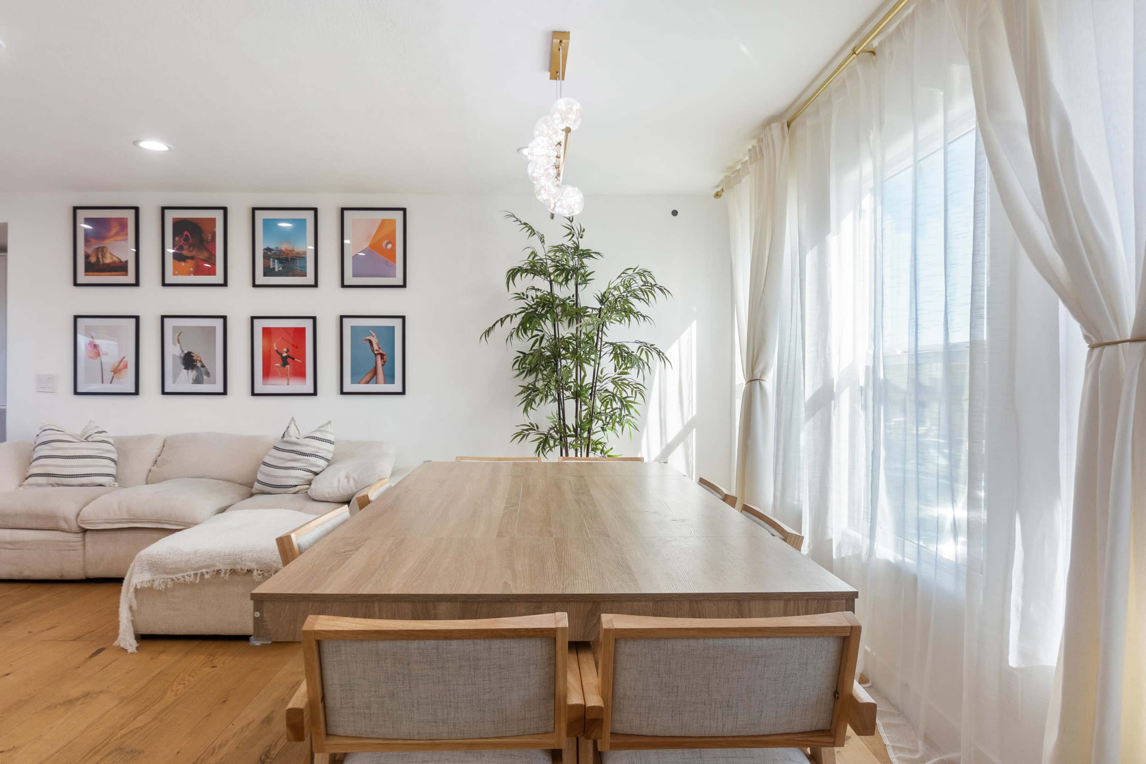 The image shows a bright dining area featuring a large wooden table with four chairs, a beige sectional sofa, and a gallery wall of framed artworks.