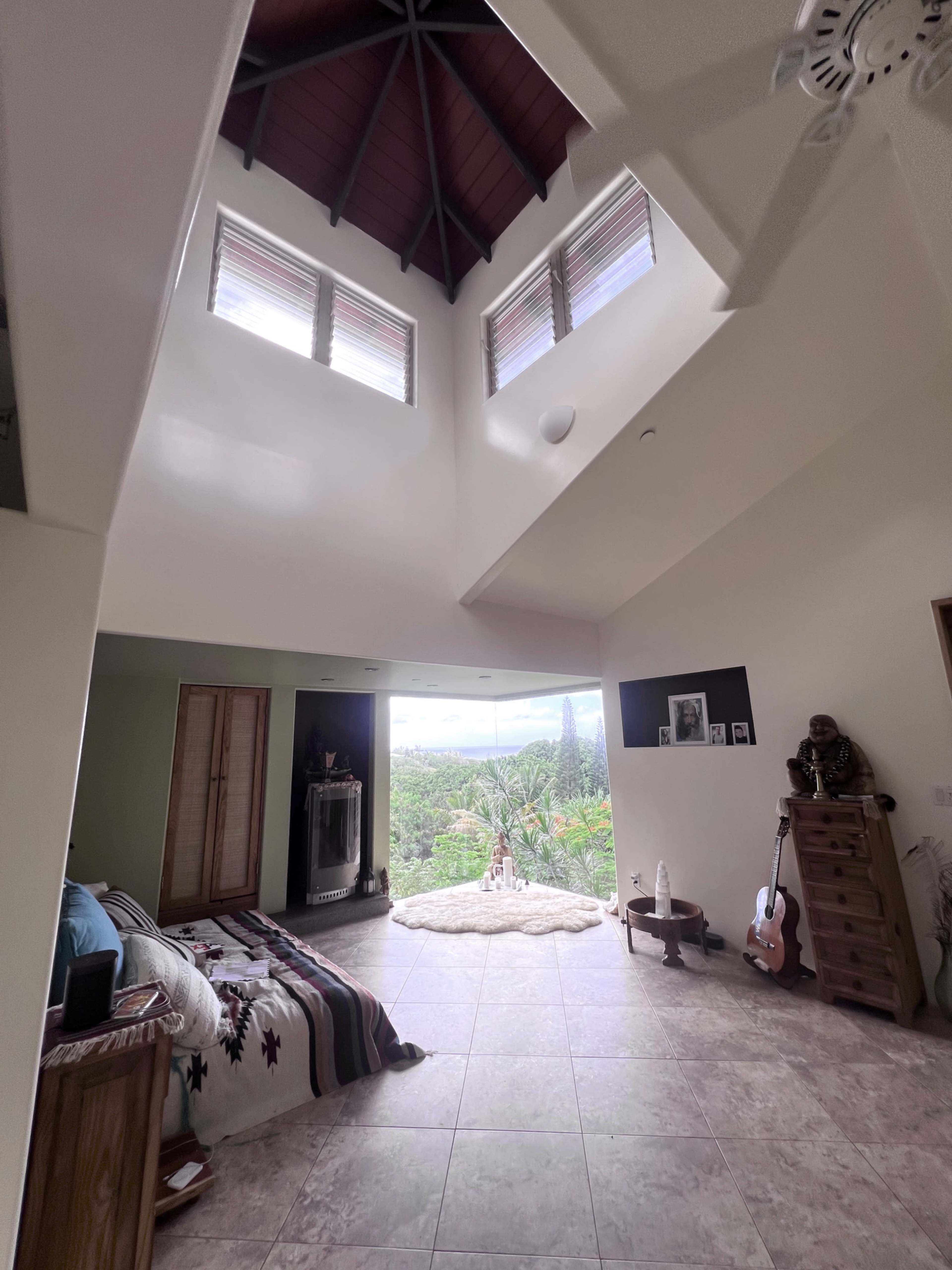 The image shows a spacious interior room with large windows providing a view of greenery, featuring a bed, wooden furniture, and decorative items.