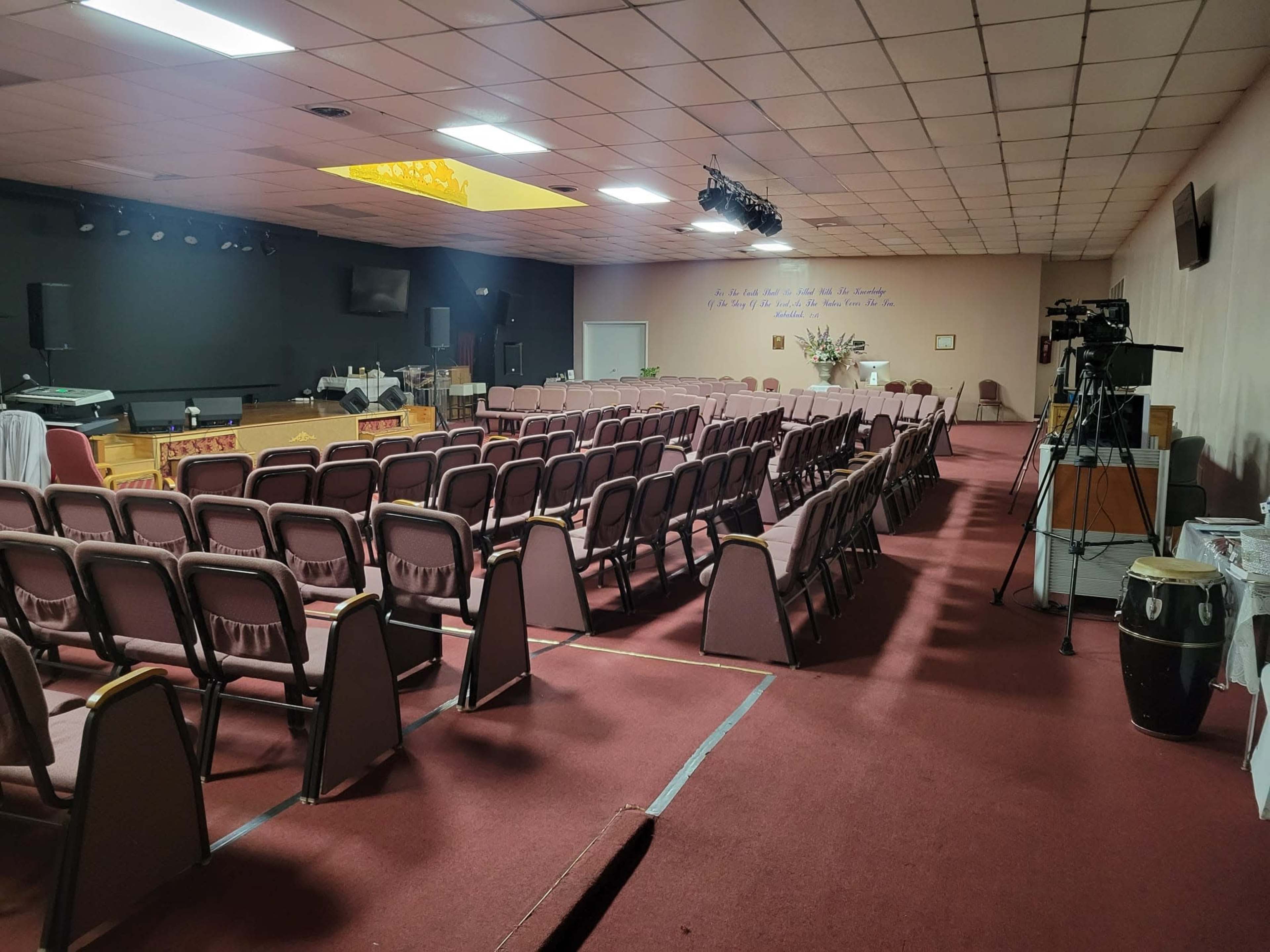 A large room set up for an event, featuring rows of empty chairs, a stage area, and a camera positioned on a tripod.