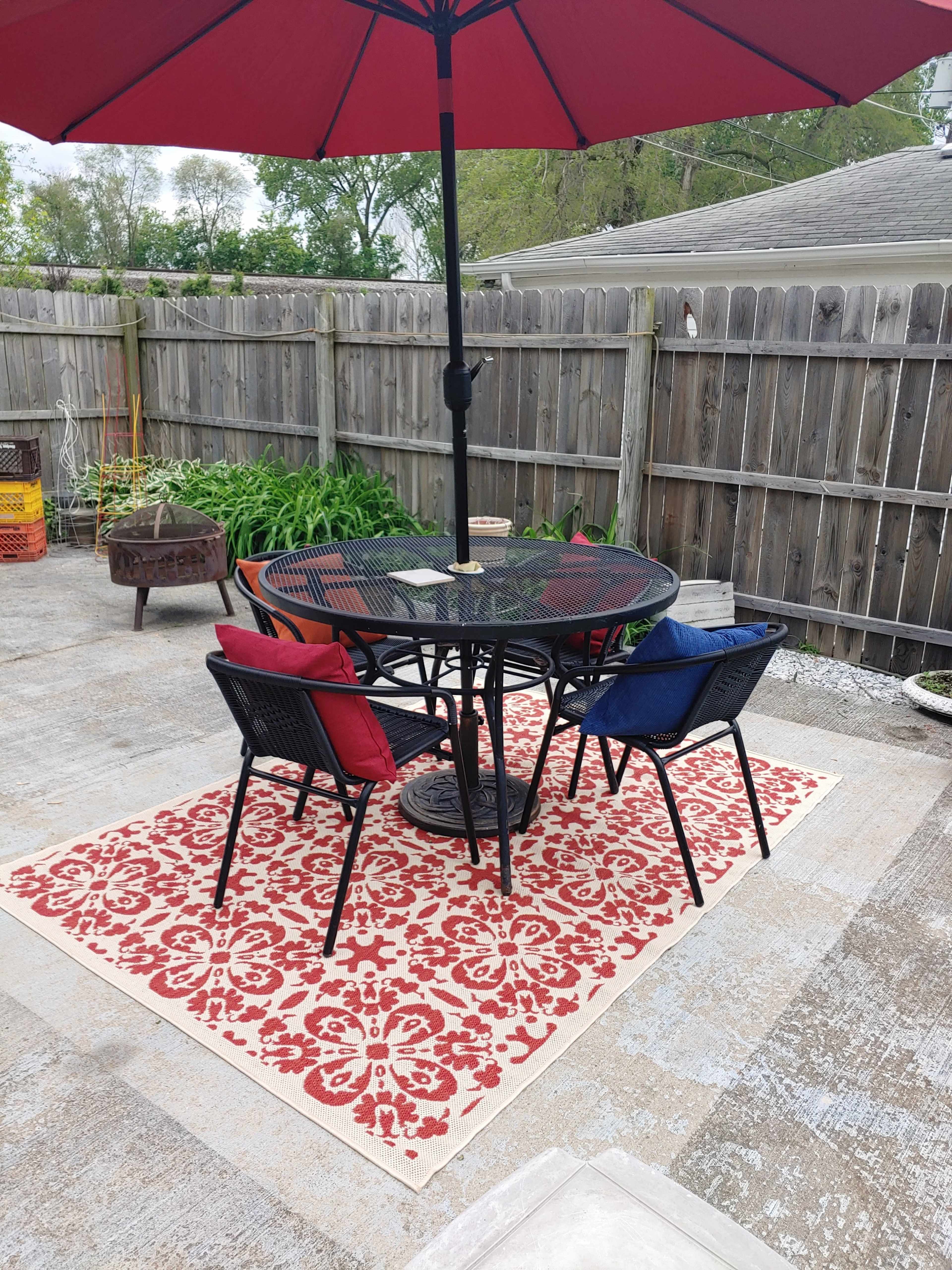A round table with two chairs is set on a patterned rug beneath a red umbrella in a fenced outdoor area.