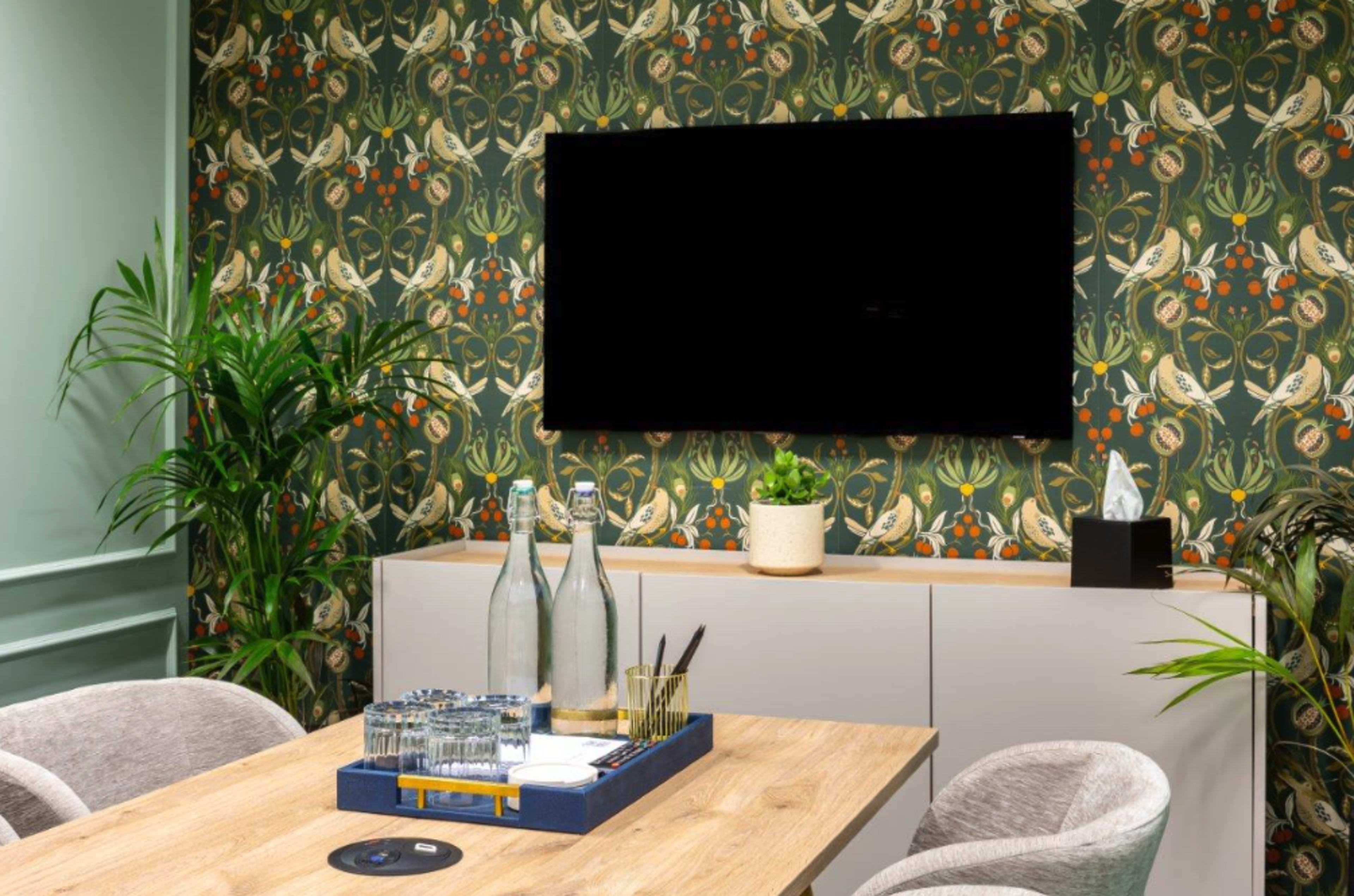 The image shows a modern interior space featuring a television mounted on a floral-patterned wall, a wooden table with drinks and supplies, and decorative plants.