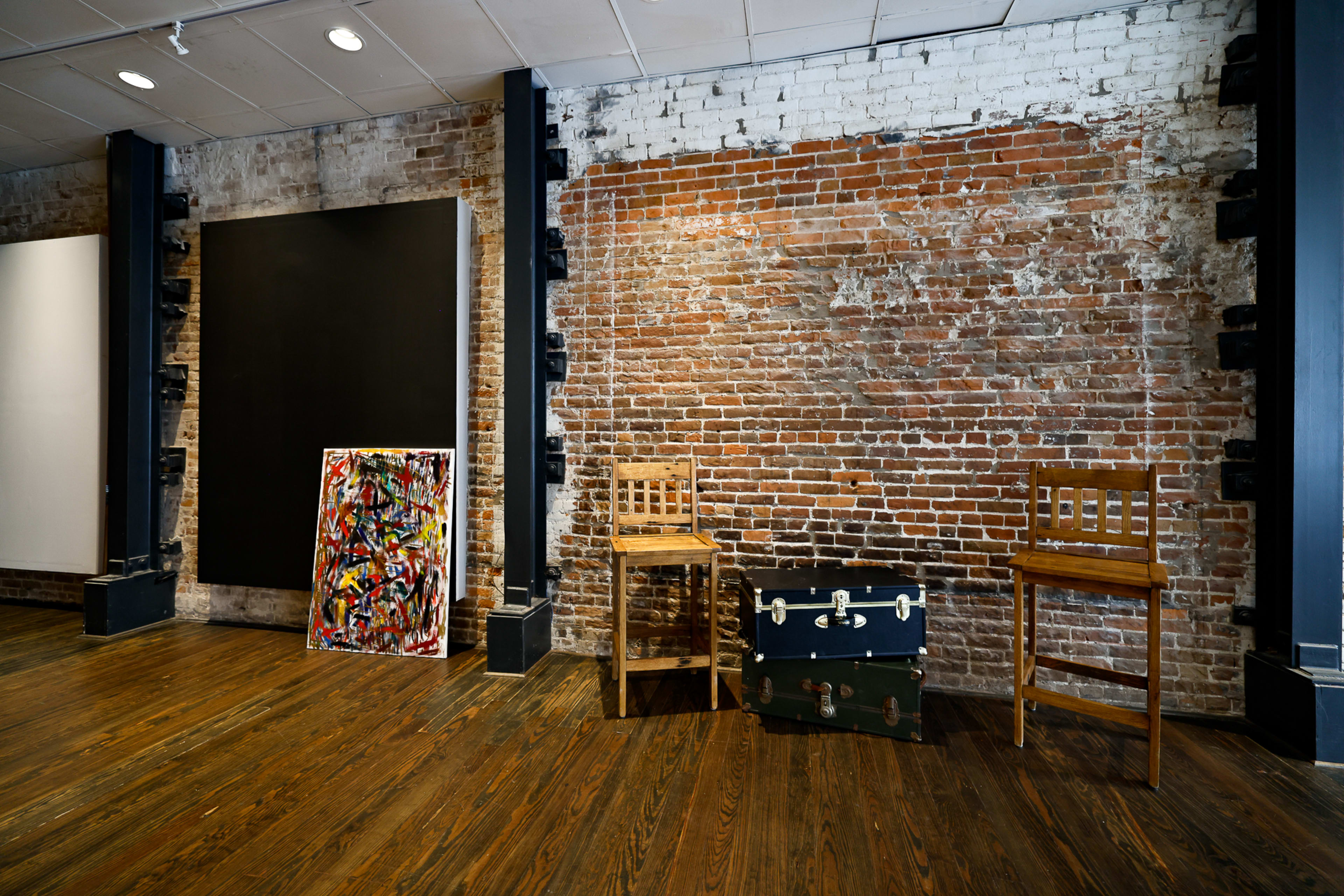 A room with exposed brick walls, wooden flooring, two wooden chairs, and a painted canvas leaning against the wall next to a vintage trunk.