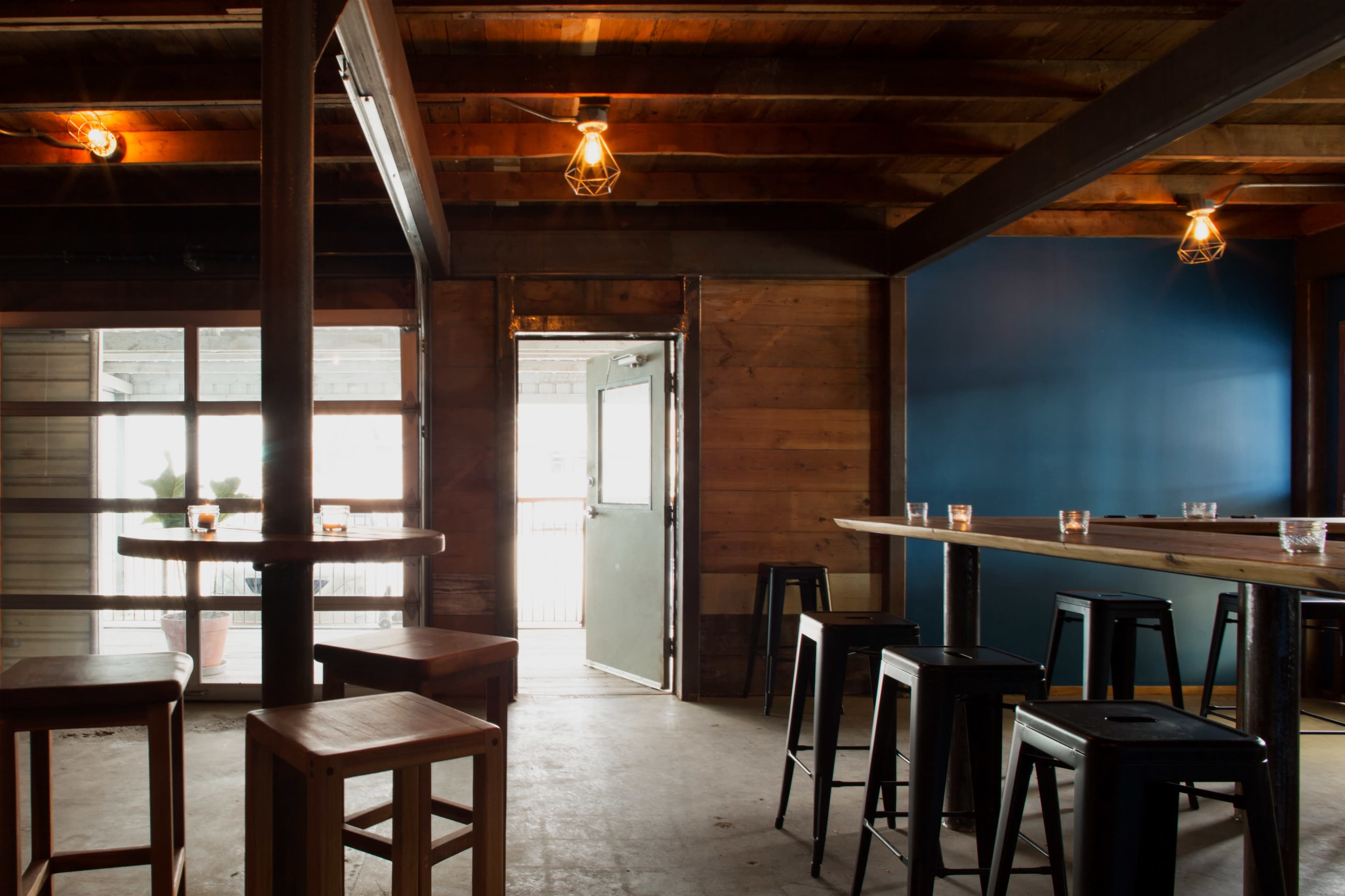 A rustic bar interior featuring wooden beams, metal stools, and an open door leading outside.