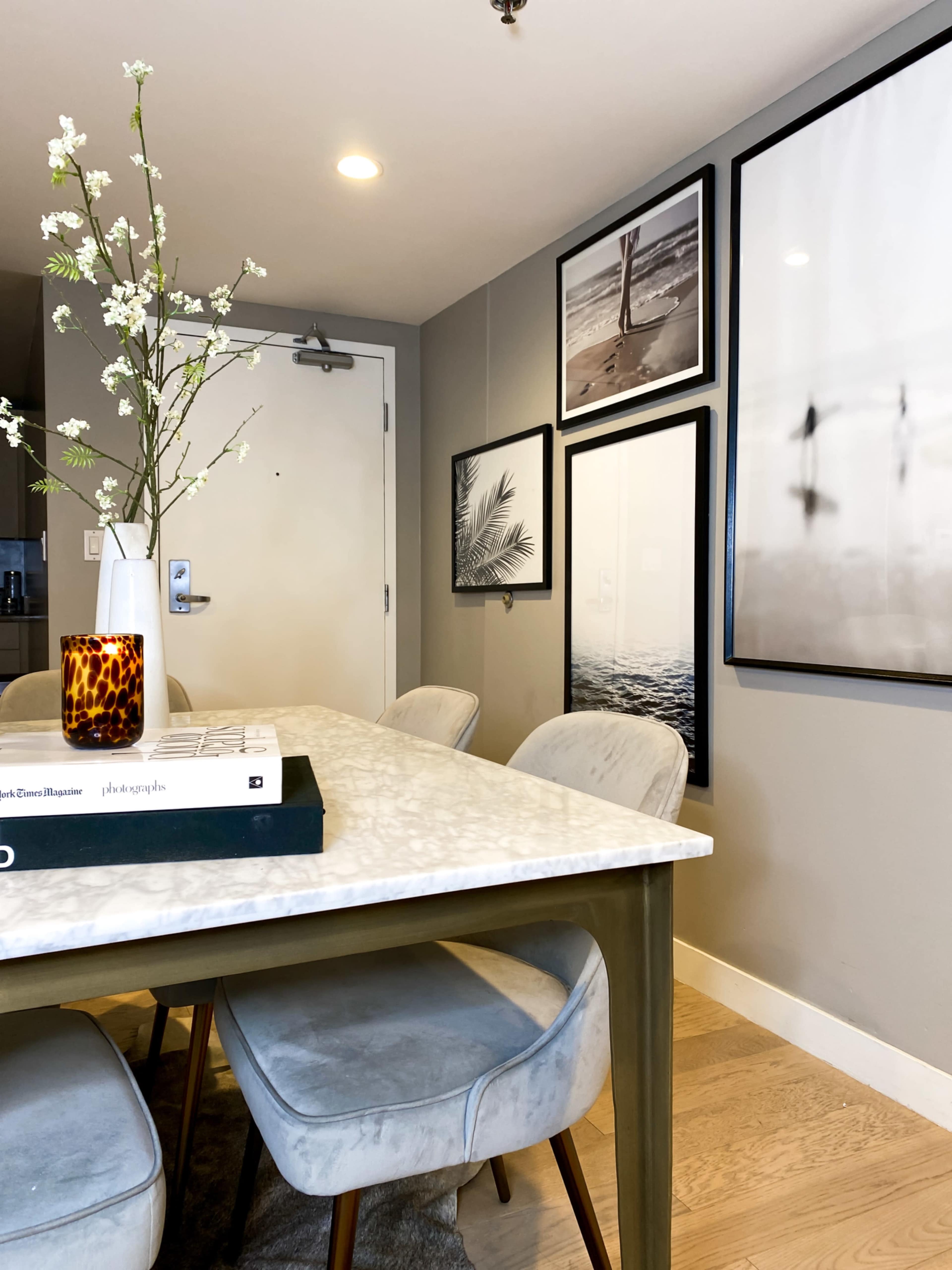 A dining area features a marble-top table surrounded by upholstered chairs, with art framed on the walls and a vase of branches as a centerpiece.