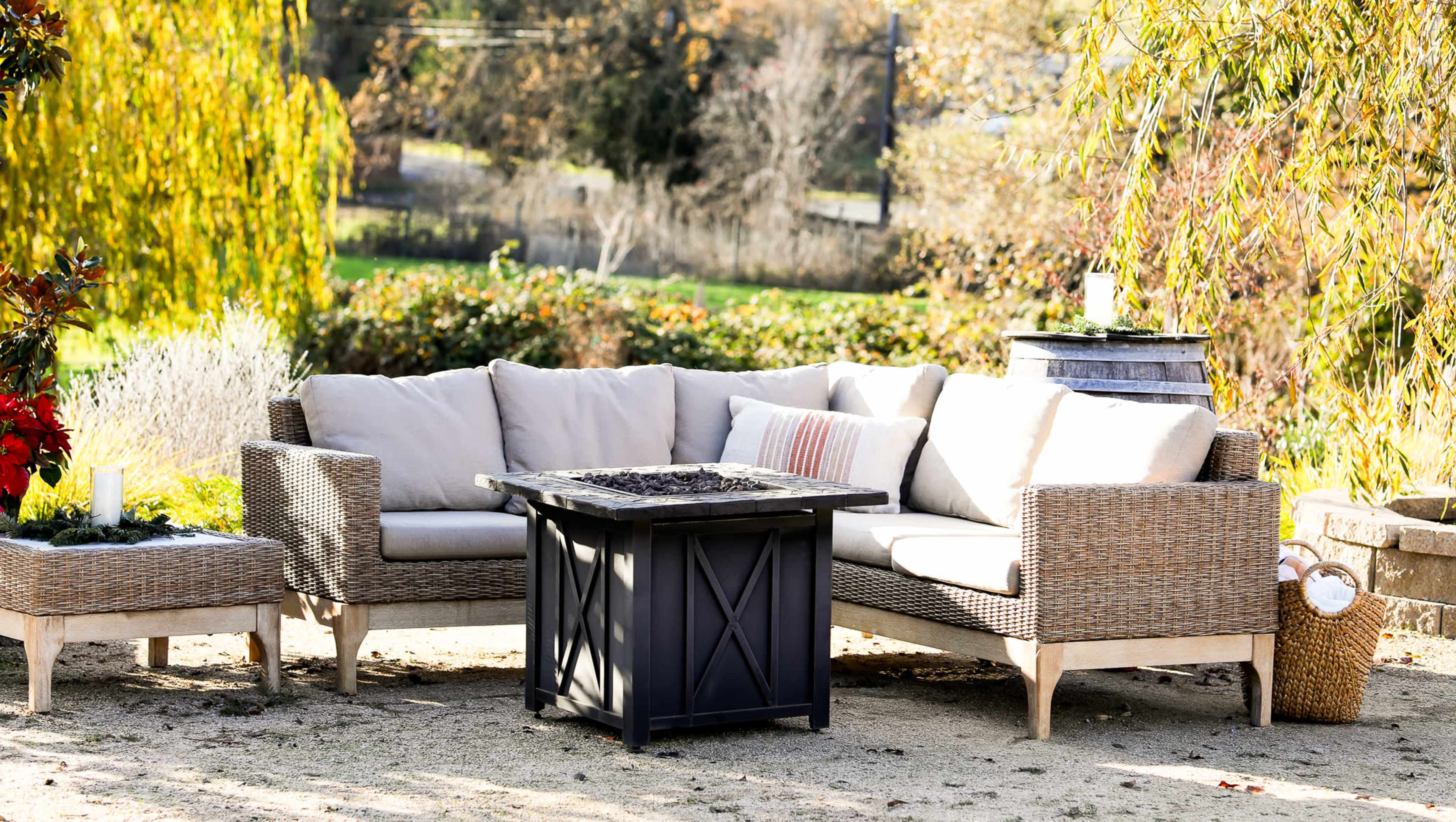 A patio seating area features a wicker sectional sofa and a black fire pit table surrounded by greenery.