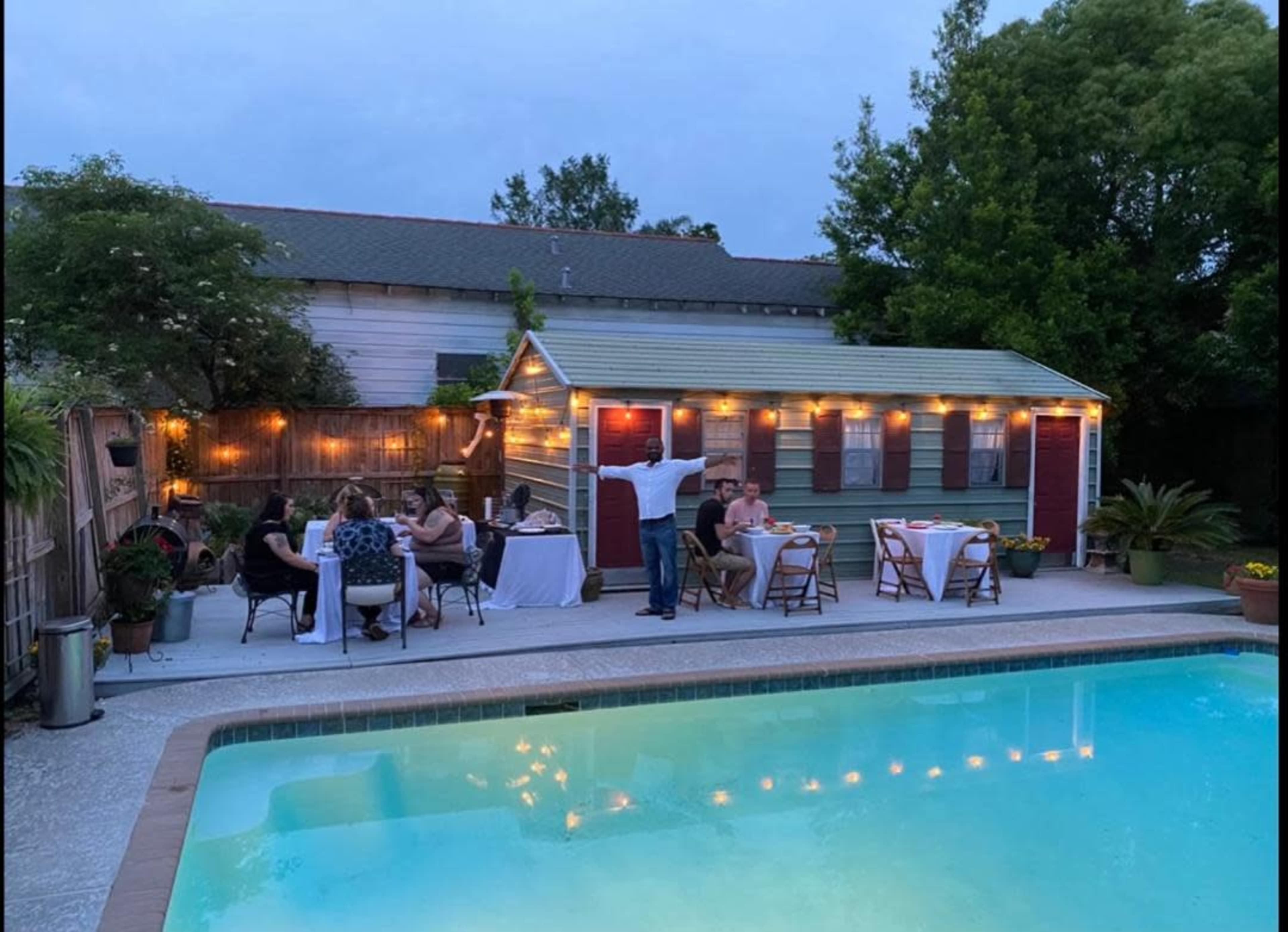 A group of people is gathered around tables near a pool at dusk, with a small building in the background adorned with string lights.