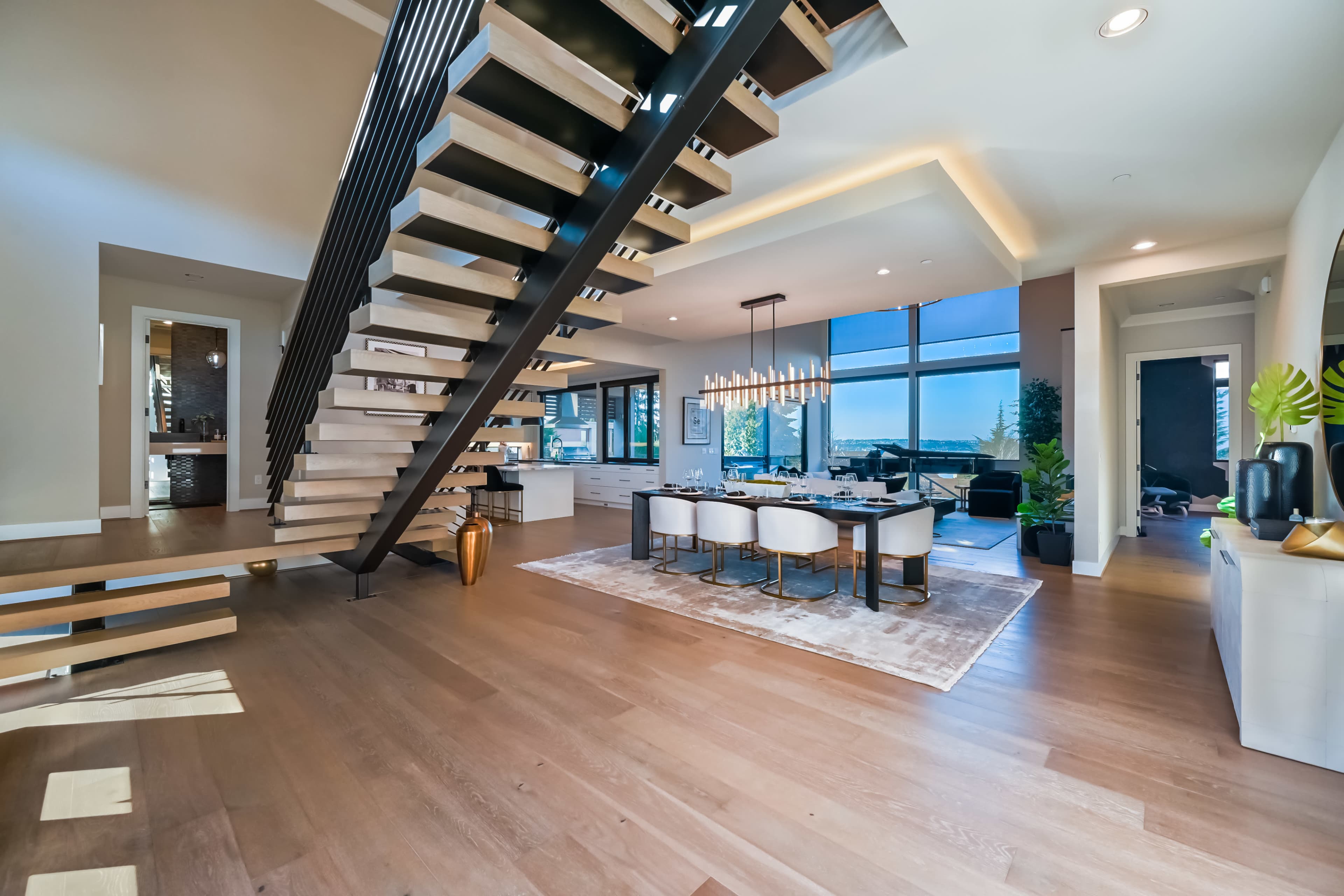 A modern interior space with an open floor plan featuring a staircase, a dining area with a table and chairs, and large windows offering a view.