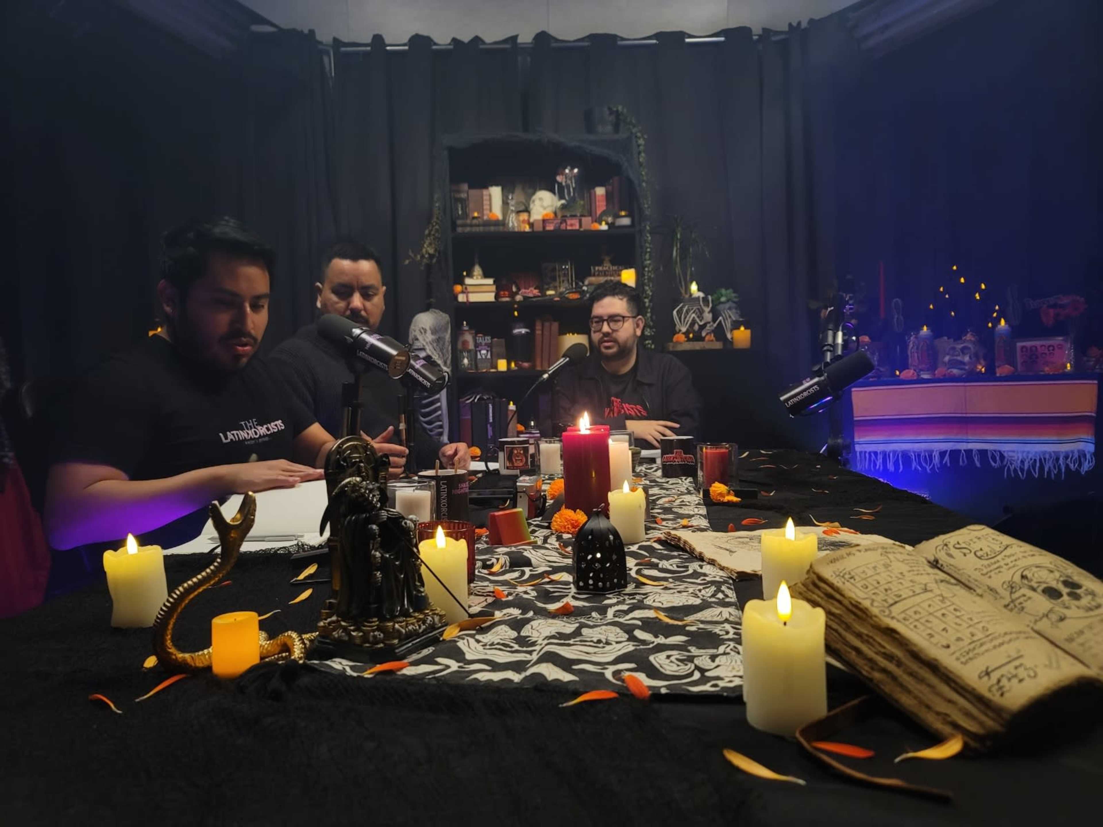 Three men sit at a table covered with candles and occult decorations, preparing for a podcast recording in a dimly lit room filled with shelves of curiosities.