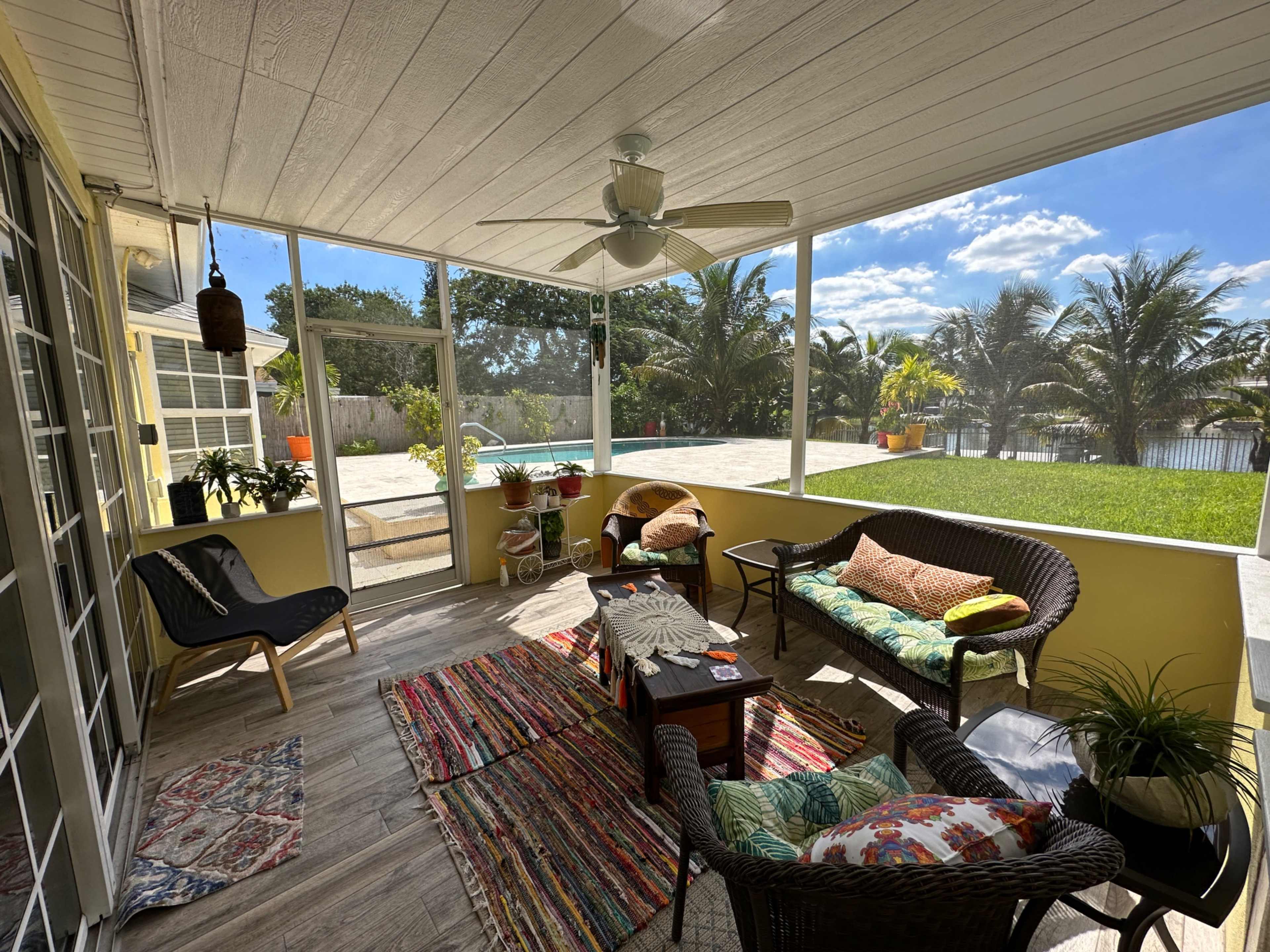 A screened porch area furnished with a coffee table, two wicker chairs, and a colorful rug, overlooking a pool and a lawn surrounded by palm trees.
